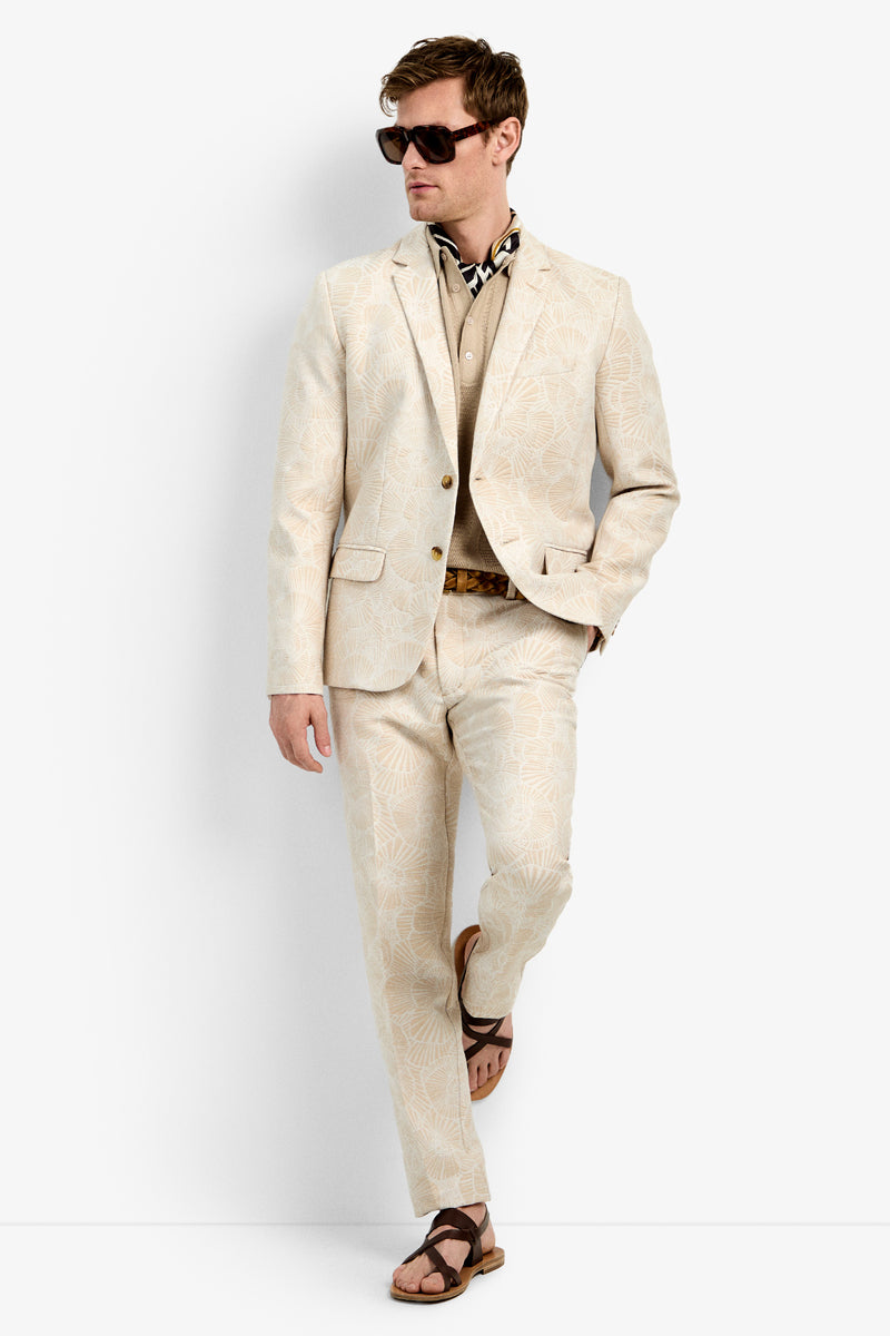 A man wearing the Thurtson Blazer, a tan shirt, brown sandals, dark sunglasses, and a patterned neck scarf stands confidently with one hand in his pocket against a plain white background.
