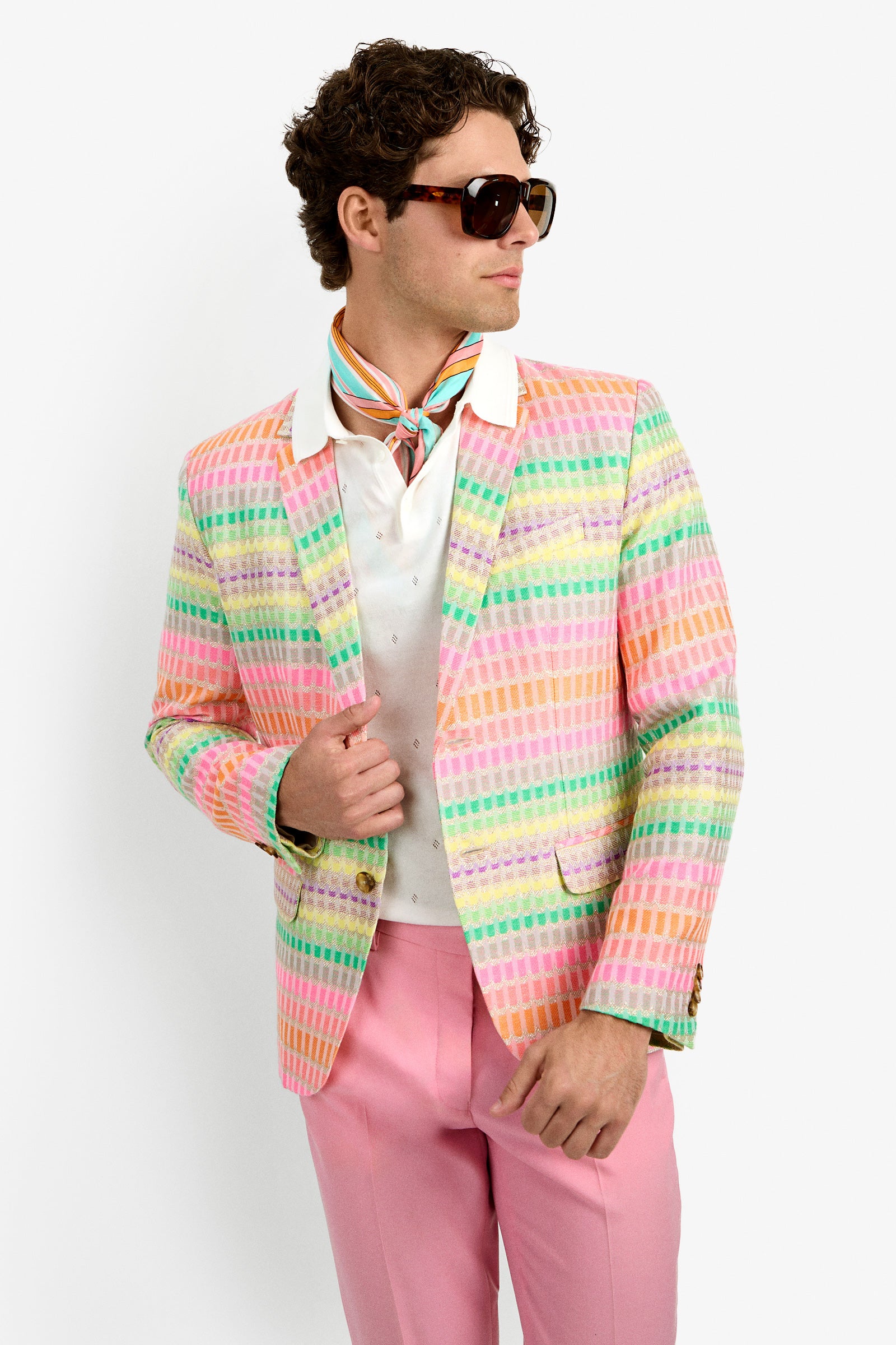 A man in the THURSTON BLAZER, featuring a pastel rainbow pattern, large sunglasses, a white shirt, colorful neck scarf, and pink pants stands against a plain white background looking off to the side.