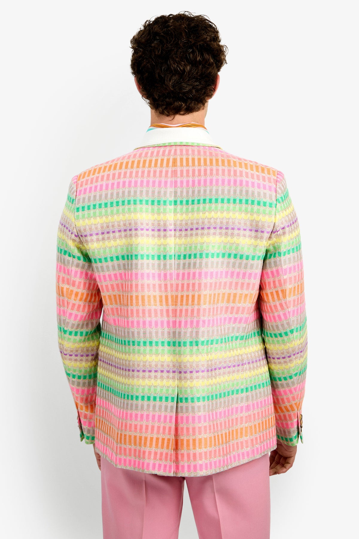 Seen from behind against a plain white background, a person with short curly hair wears the Thurston Blazer—a colorful pastel plaid jacket in pink, orange, yellow, green, and blue—paired with light pink pants.