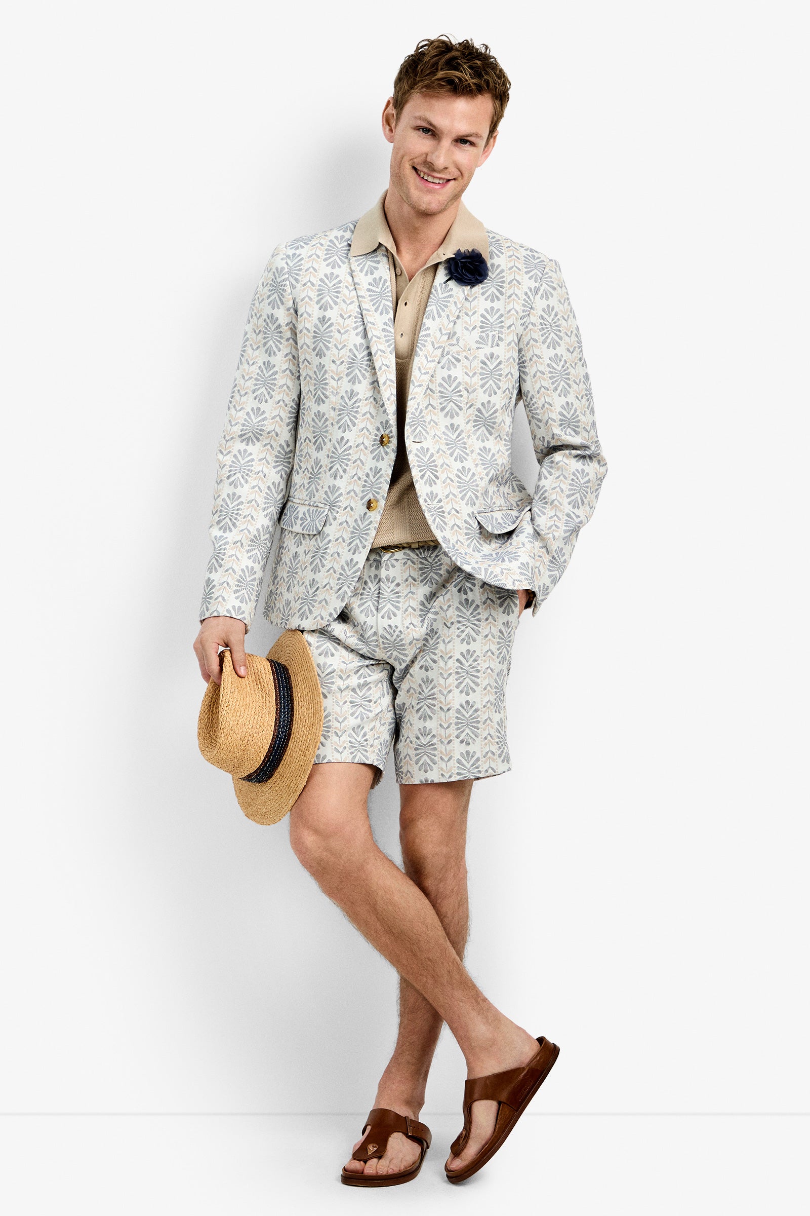 A man smiles in the Thurston Blazer with matching shorts, a beige polo shirt, and brown sandals, holding a straw hat. He poses casually against a plain white background.