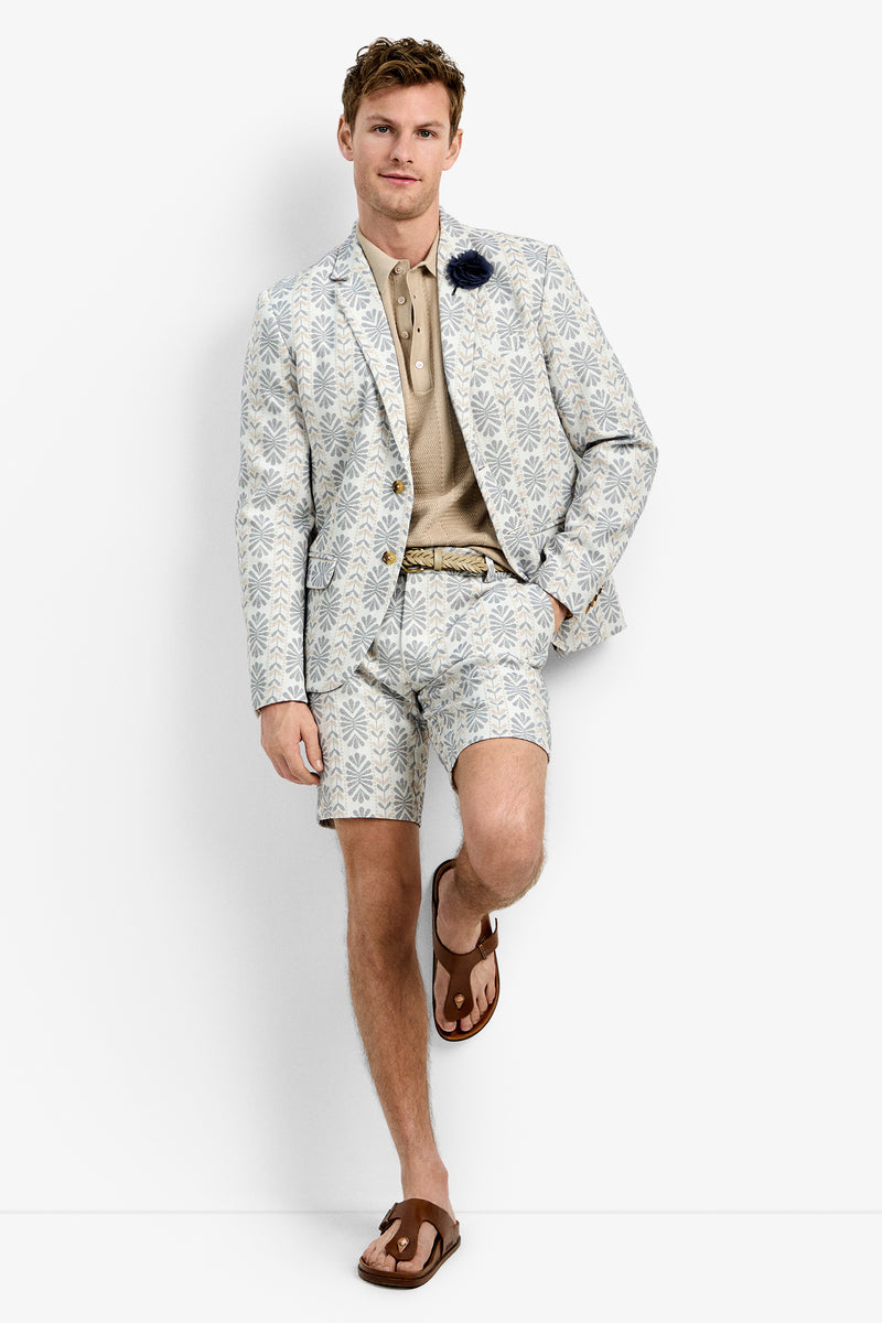 A man stands relaxed with one leg bent against a white background, wearing the Thurston Blazer—a light-colored, patterned suit jacket—matched with shorts, a beige polo shirt, brown sandals, and a dark flower lapel pin.