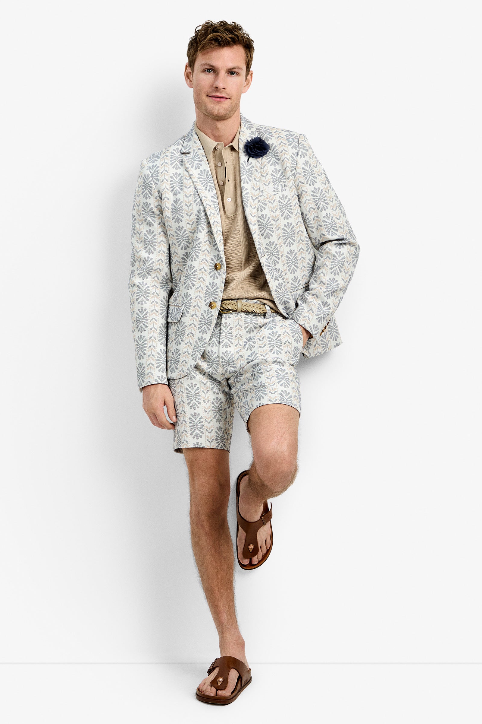 A man stands relaxed with one leg bent against a white background, wearing the Thurston Blazer—a light-colored, patterned suit jacket—matched with shorts, a beige polo shirt, brown sandals, and a dark flower lapel pin.