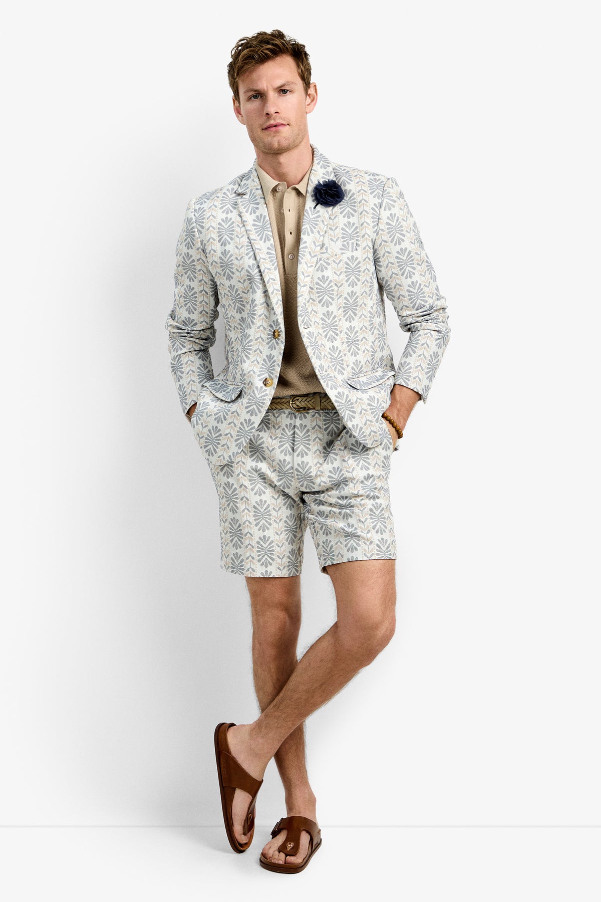 A man with short hair stands against a white background wearing the Thurston Blazer and matching shorts, a beige polo shirt, brown sandals, and a dark flower lapel pin, with his hands in his pockets.
