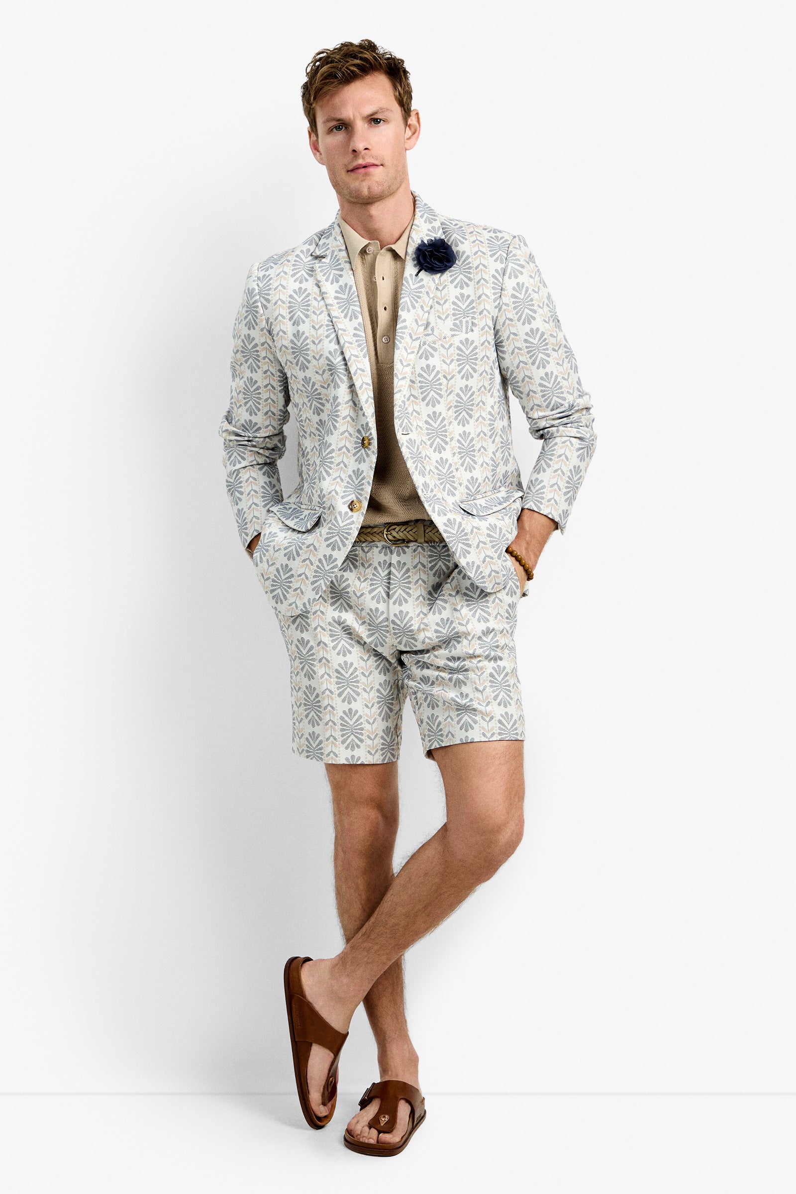 A man with short hair stands against a white background wearing the Thurston Blazer and matching shorts, a beige polo shirt, brown sandals, and a dark flower lapel pin, with his hands in his pockets.