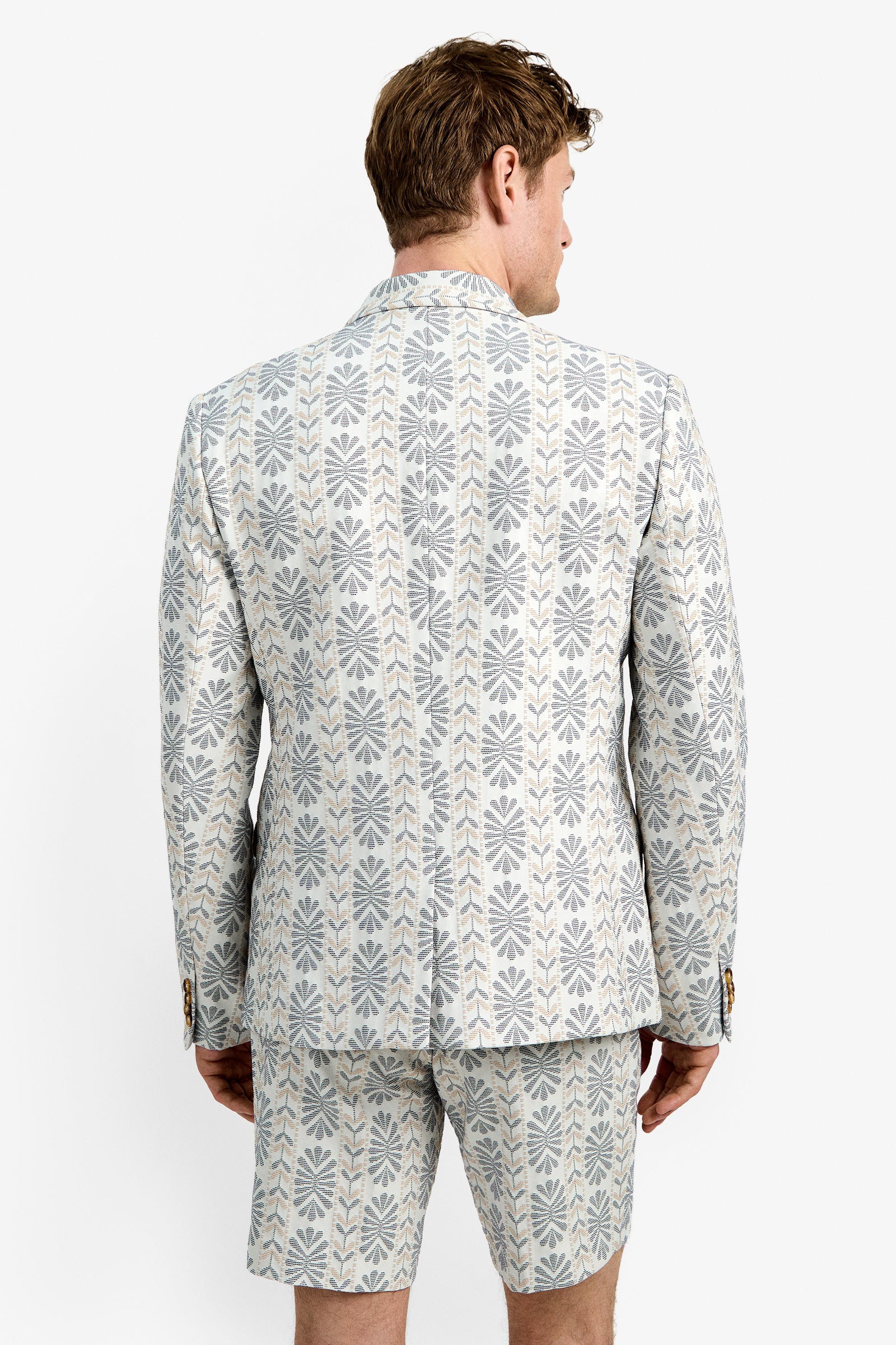 A man stands with his back to the camera, wearing the Thurston Blazer and matching shorts, both featuring a repeating geometric floral pattern in pale gray and beige.