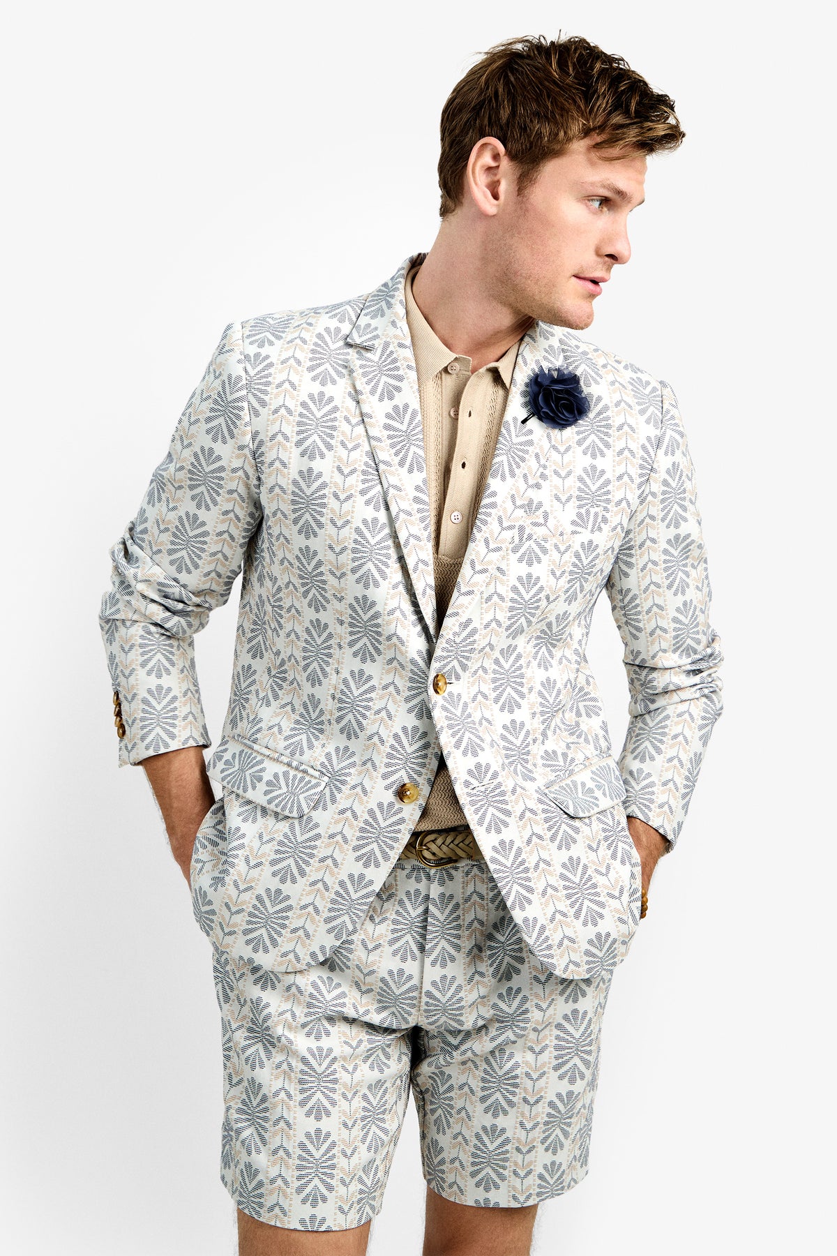 A man stands with hands in pockets, looking to the side against a plain background, wearing the Thurston Blazer and matching shorts over a beige shirt, accessorized with a dark flower pin on the lapel.
