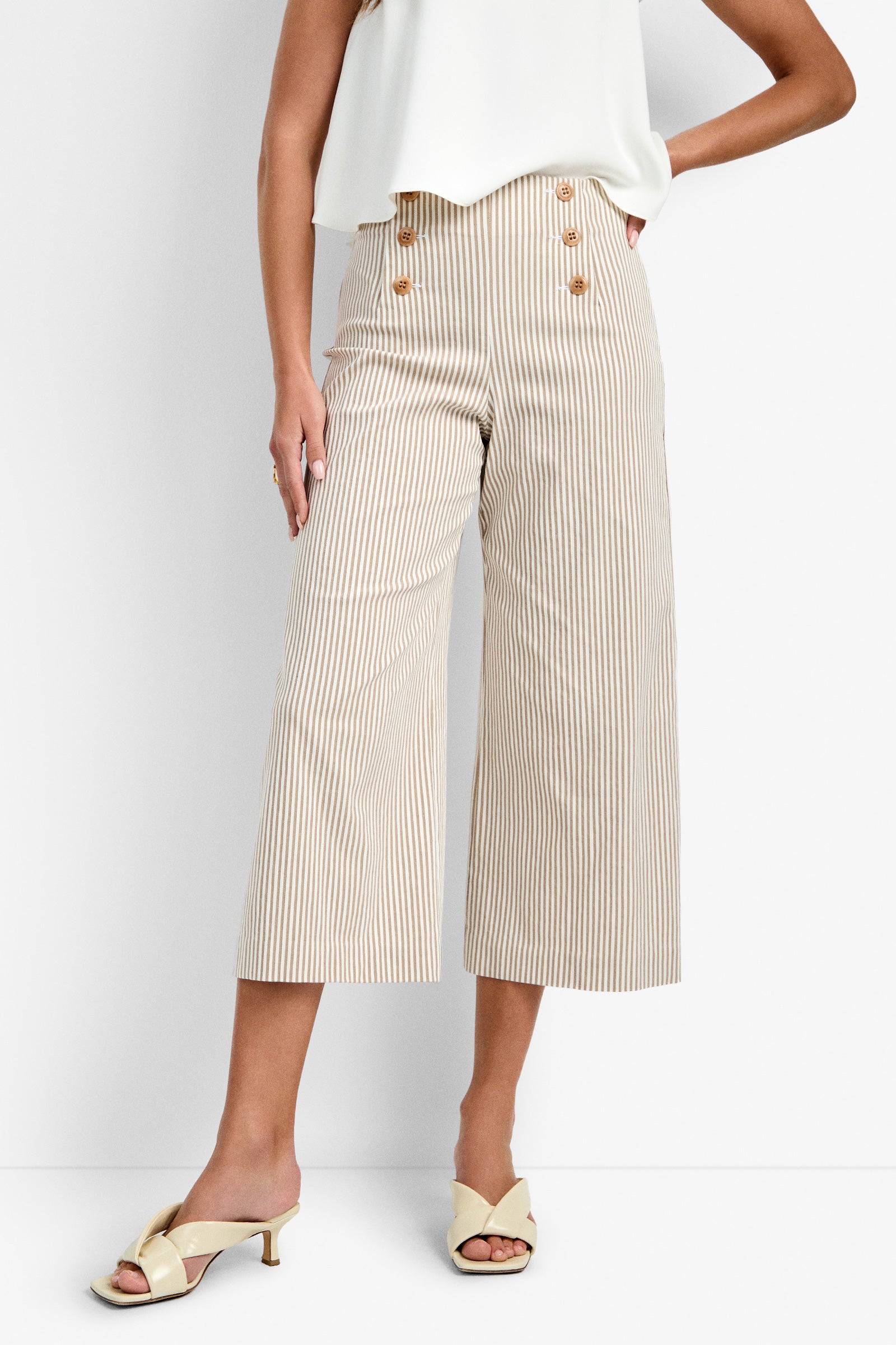 A woman wears the Saltie Pant—cream and beige vertical stripes, cropped with large front buttons—paired with a sleeveless white top and cream heeled sandals, standing against a white background.