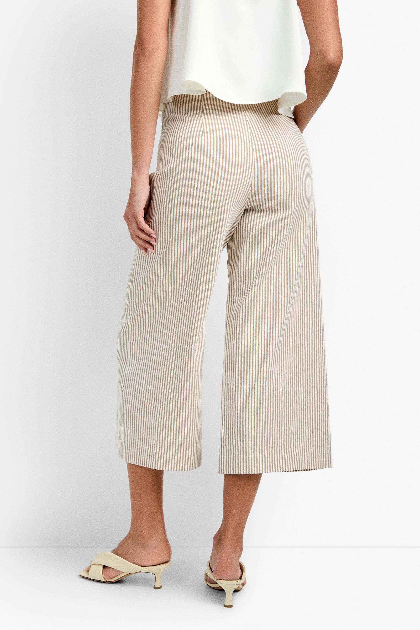 A person wearing the Saltie Pant—beige and white vertically striped cropped wide-leg pants—with a white sleeveless top and cream heeled mules, stands with their back to the camera against a plain white background.