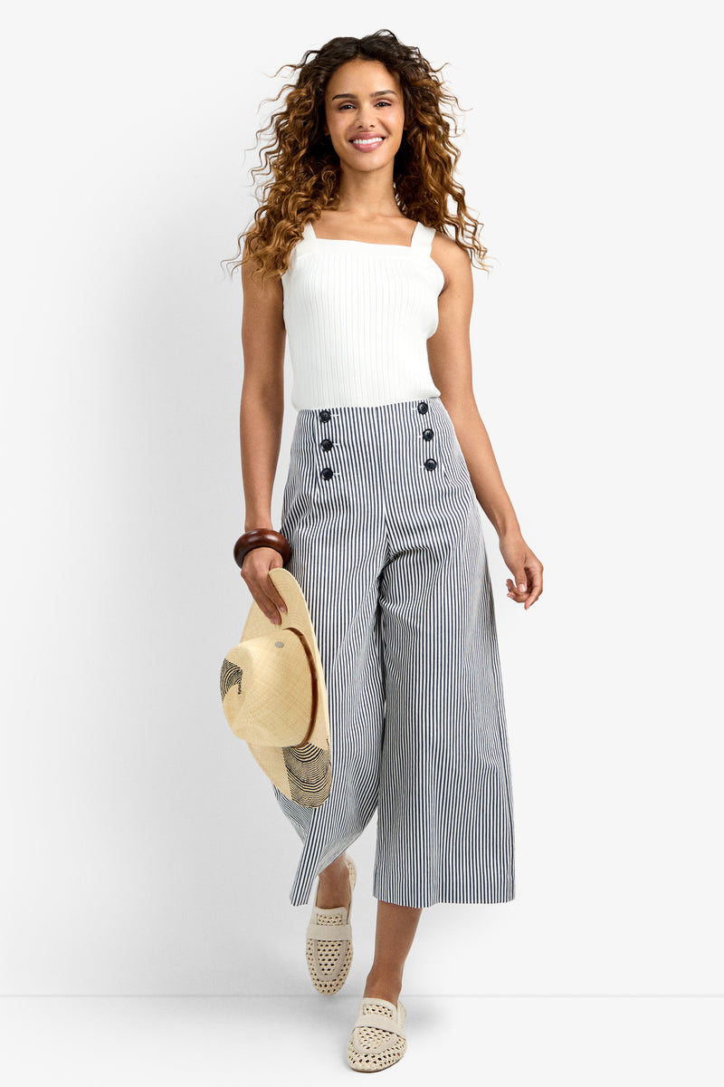 A woman with curly hair smiles while wearing a white sleeveless top, Saltie Pant—high-waisted striped culottes with buttons—white woven slip-on shoes, and holding a straw hat against a plain white background.