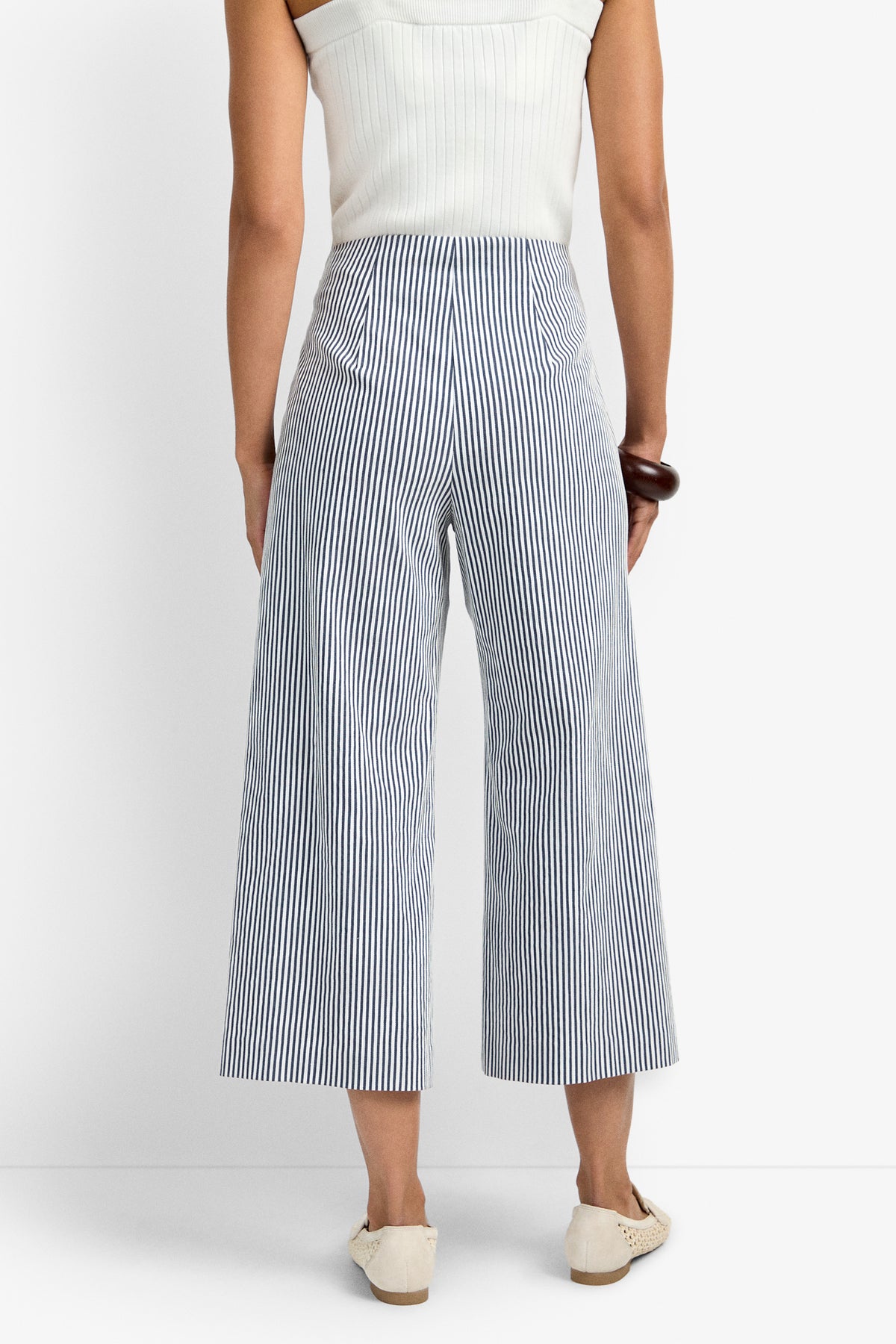 A person is seen from the back wearing a sleeveless white top and the Saltie Pant—high-waisted, wide-leg blue-and-white vertical striped pants with two back pockets—paired with white shoes.