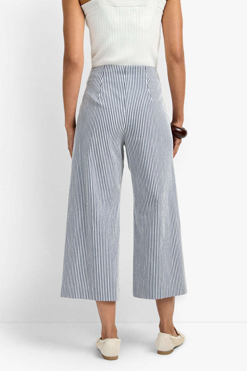 A person is seen from the back wearing a sleeveless white top and the Saltie Pant—high-waisted, wide-leg blue-and-white vertical striped pants with two back pockets—paired with white shoes.