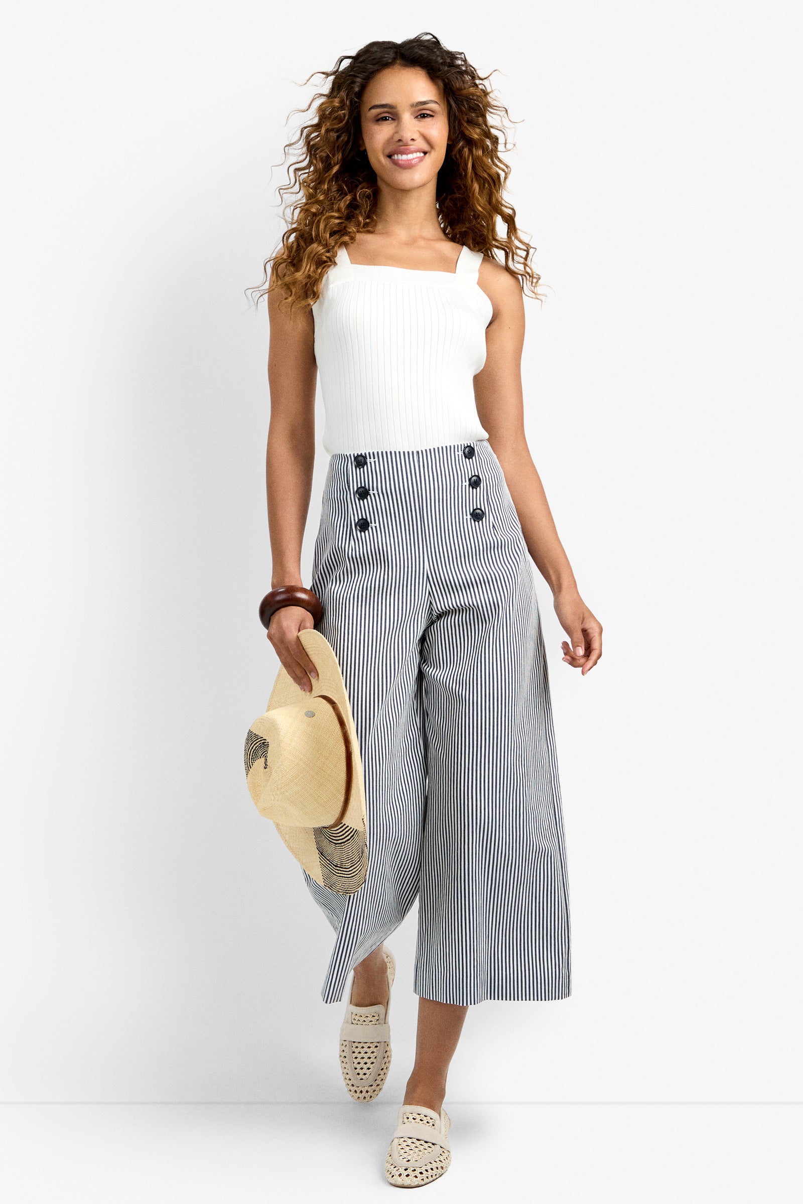 A woman with curly hair wears the Saltie Pant, a pair of high-waisted, wide-leg striped pants with buttons, paired with a white sleeveless top. She holds a straw hat and wears woven slip-on shoes, smiling as she walks.