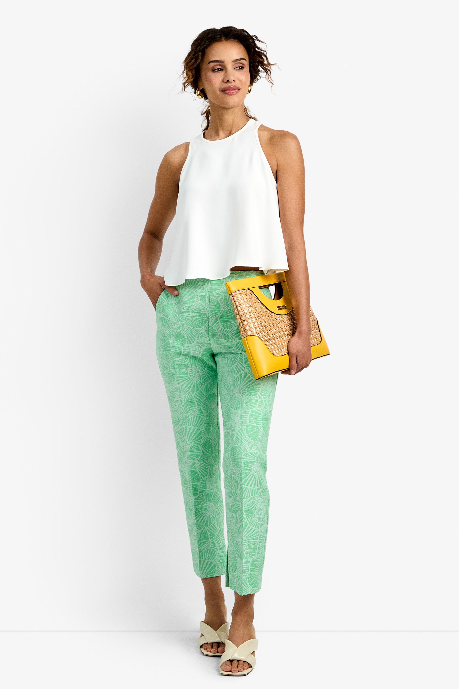 A woman stands against a white background wearing a sleeveless white top, Pop Pant in Lush Lagoon, white sandals, and holding a yellow woven clutch. She smiles with one hand in her pocket.