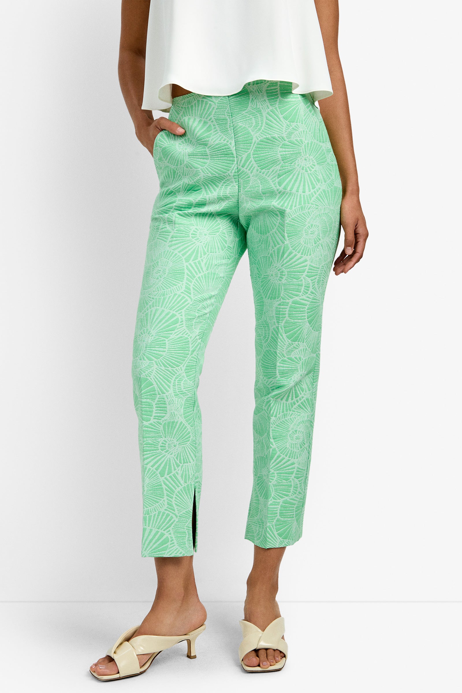 A person wearing the Pop Pant in light green with a white floral pattern, paired with a white top and cream heeled sandals, standing with hands in pockets.