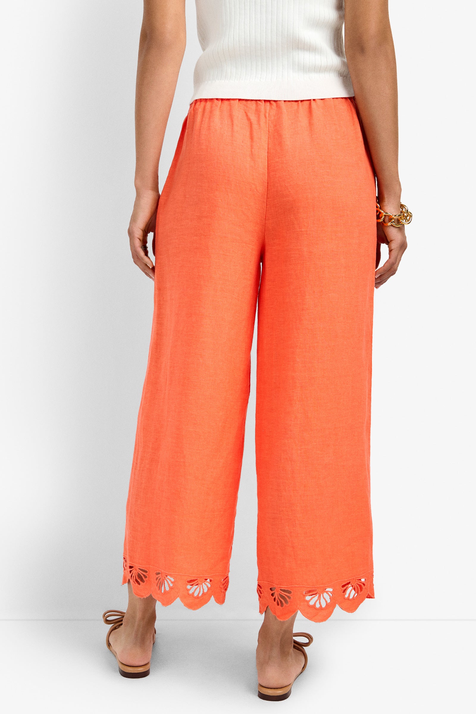 A person stands facing away, wearing the Cruze Pant in bright orange with scalloped cut-out hems, paired with a white ribbed top and tan sandals.