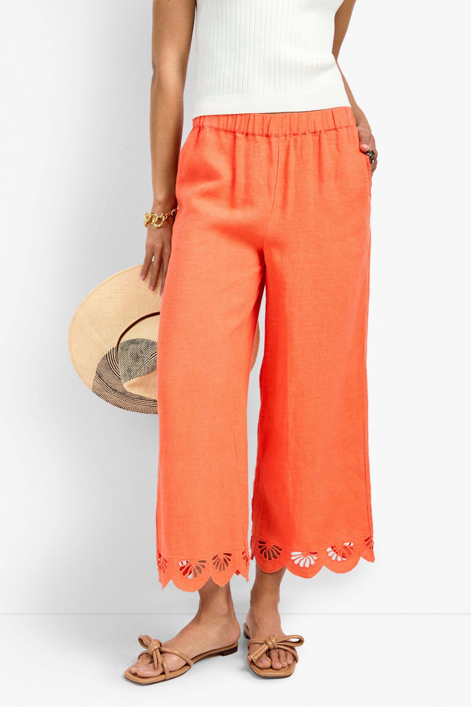 Someone wearing the Cruze Pant in coral with scalloped, laser-cut hems, paired with a white sleeveless top, tan bow sandals, a gold bracelet, and holding a straw hat for a casual, summery look.