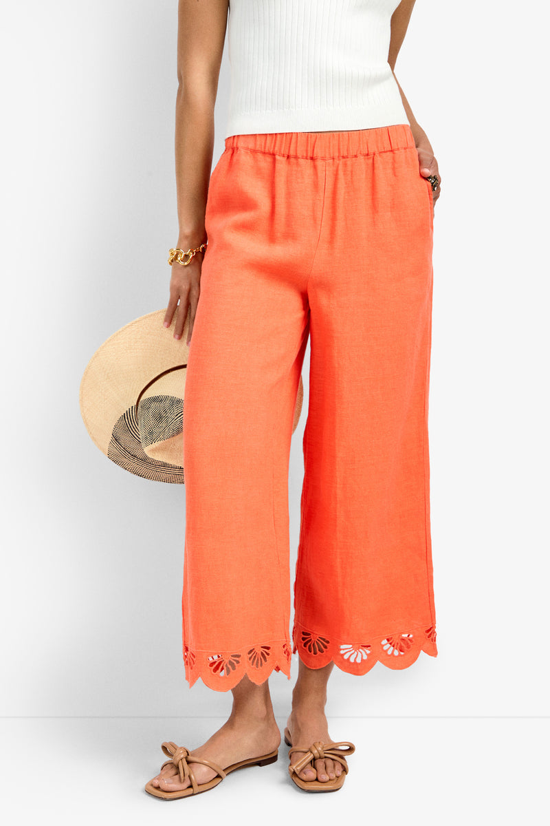 Someone wearing the Cruze Pant in coral with scalloped, laser-cut hems, paired with a white sleeveless top, tan bow sandals, a gold bracelet, and holding a straw hat for a casual, summery look.