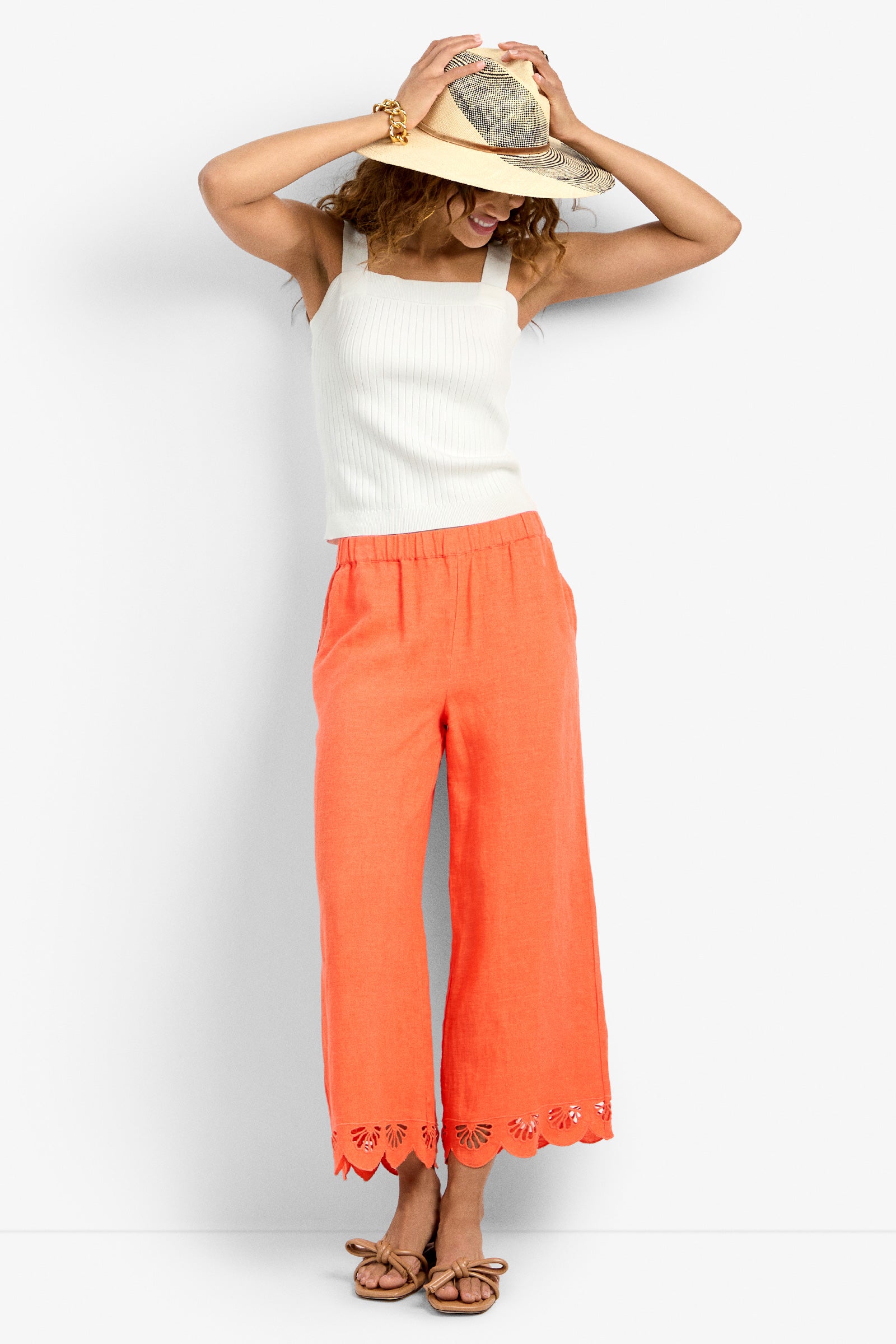 A woman in the Cruze Pant—coral, wide-leg with cut-out hem details—paired with a white sleeveless top, brown sandals, and a straw hat smiles, one hand holding her hat and the other on her head.