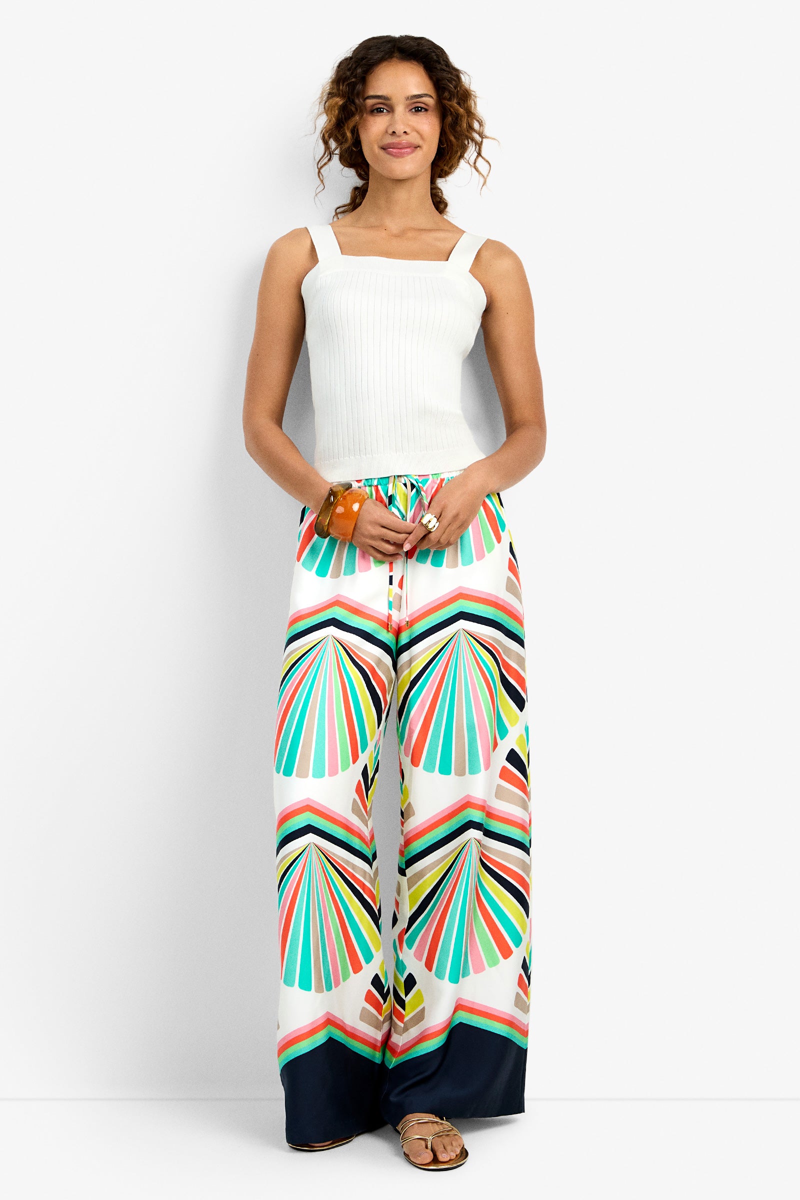 A woman with curly hair smiles, wearing a white sleeveless ribbed top and the Dellmar Pant in MULTI, which features a colorful geometric pattern. She accessorizes with a bracelet and sandals, posing against a plain white background.