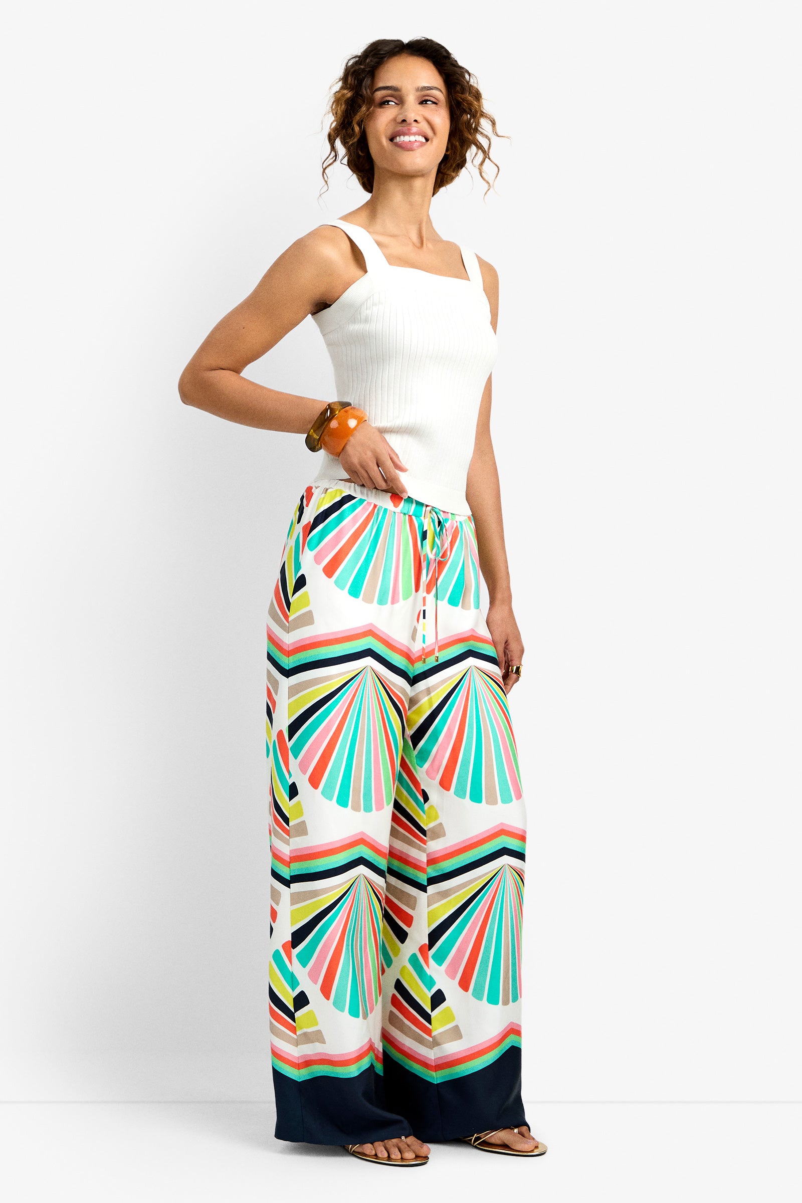 A woman smiles in the Dellmar Pant, featuring a colorful geometric fan pattern, paired with a white sleeveless top, bangle, and sandals. She poses against a plain white background.
