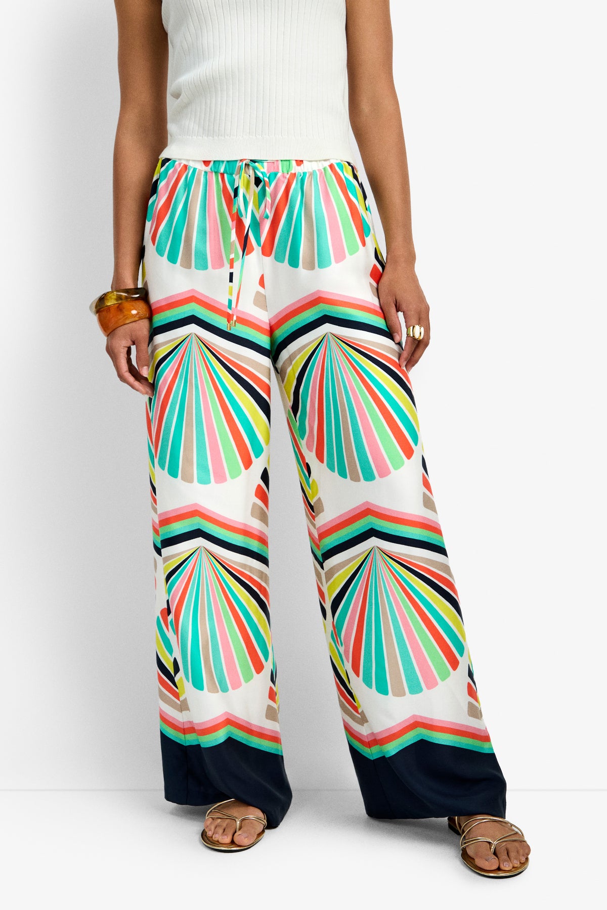 A person wearing the Dellmar Pant, featuring a colorful scallop shell pattern in teal, yellow, orange, pink, and black, styled with a white sleeveless top, brown bangle, and flat sandals.