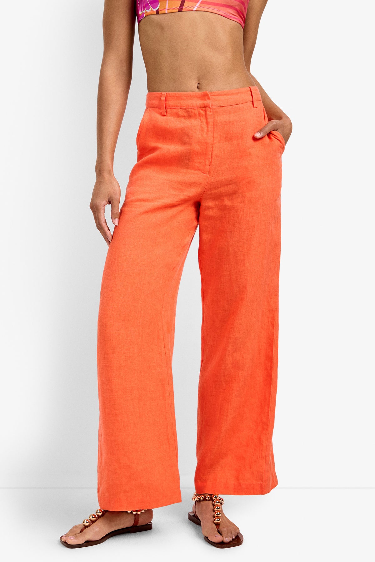 A person wearing CALYPSO CORAL Tonga Pant wide-leg linen pants, a pink and orange patterned crop top, and brown sandals with beaded ankle straps stands against a plain white background.