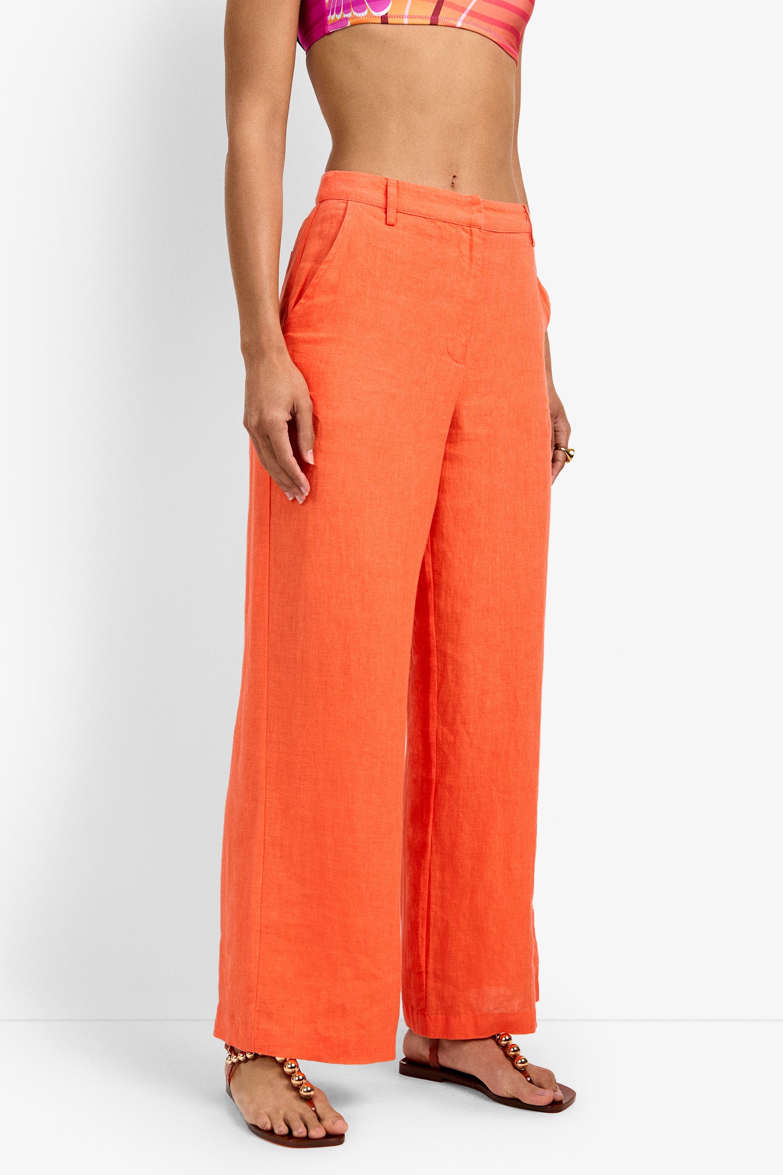 A person wearing high-waisted, wide-leg orange Tonga Pants with a colorful crop top and flat sandals stands against a plain white background. The image is cropped above the shoulders.