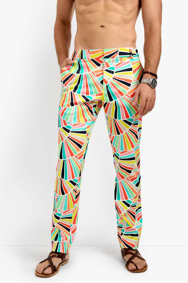 A man models the Clyde Slim Trouser, featuring a vibrant geometric fan-like pattern in yellow, green, orange, pink, and black. He pairs the pants with sandals and a watch, standing shirtless with one hand in his pocket.