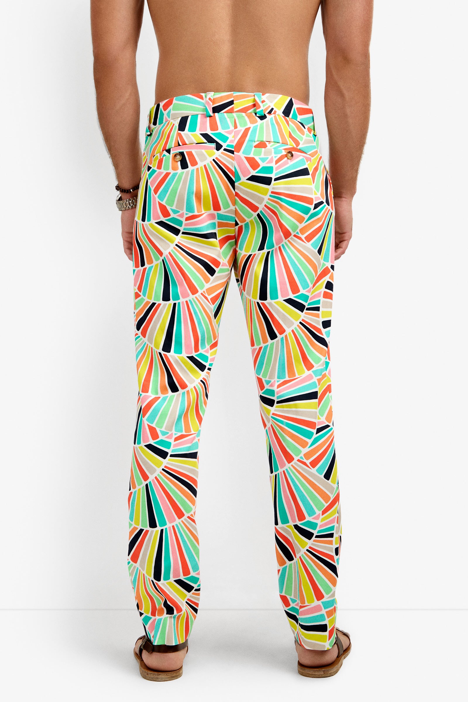 A shirtless person faces away, wearing Clyde Slim Trouser featuring a fan-like geometric pattern in orange, green, yellow, black, and white. They also wear sandals and a bracelet.