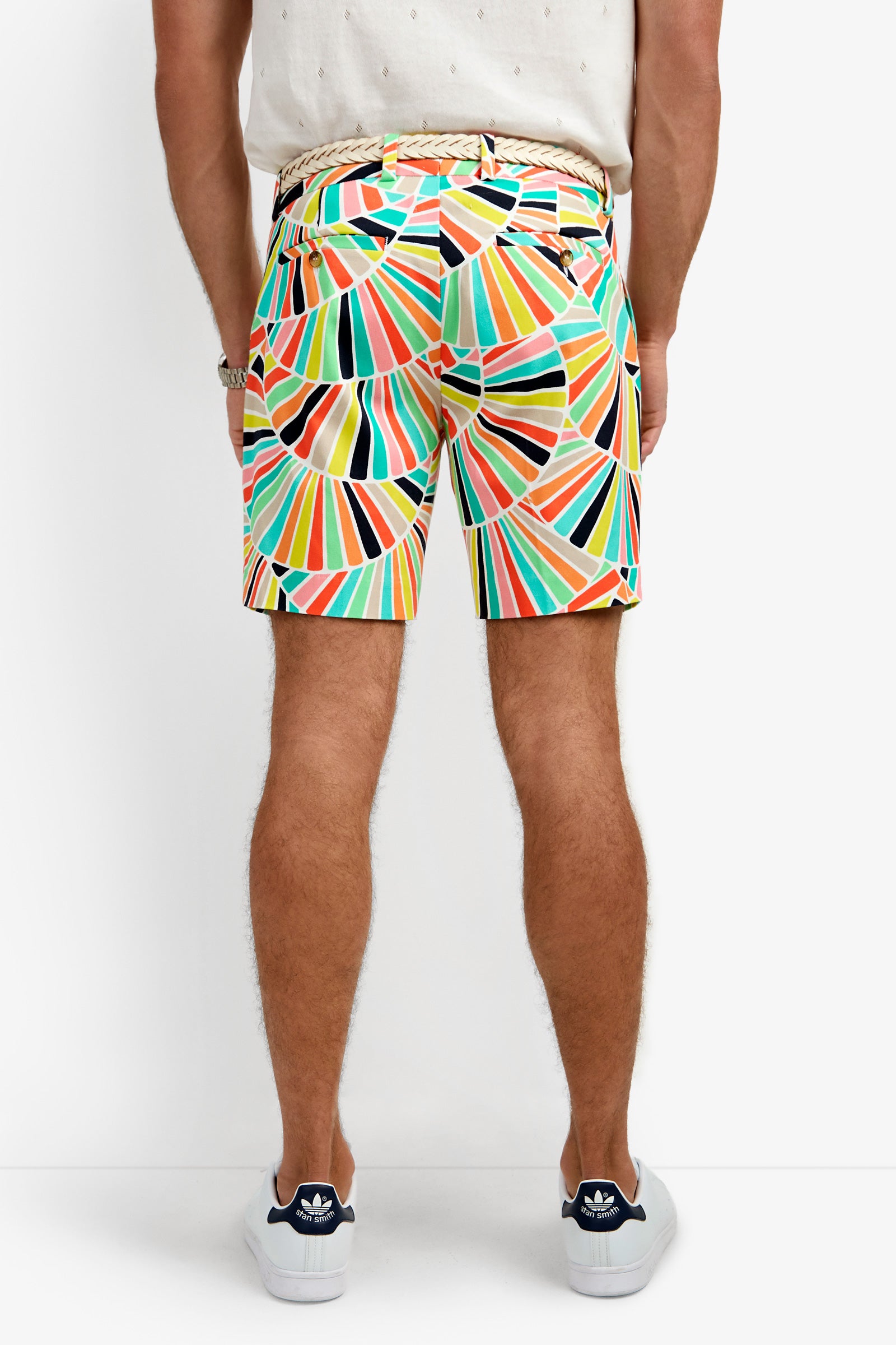 A person faces away, wearing the Lawrence Short with a colorful geometric pattern, a white shirt, beige woven belt, and white sneakers with navy accents against a plain white background.
