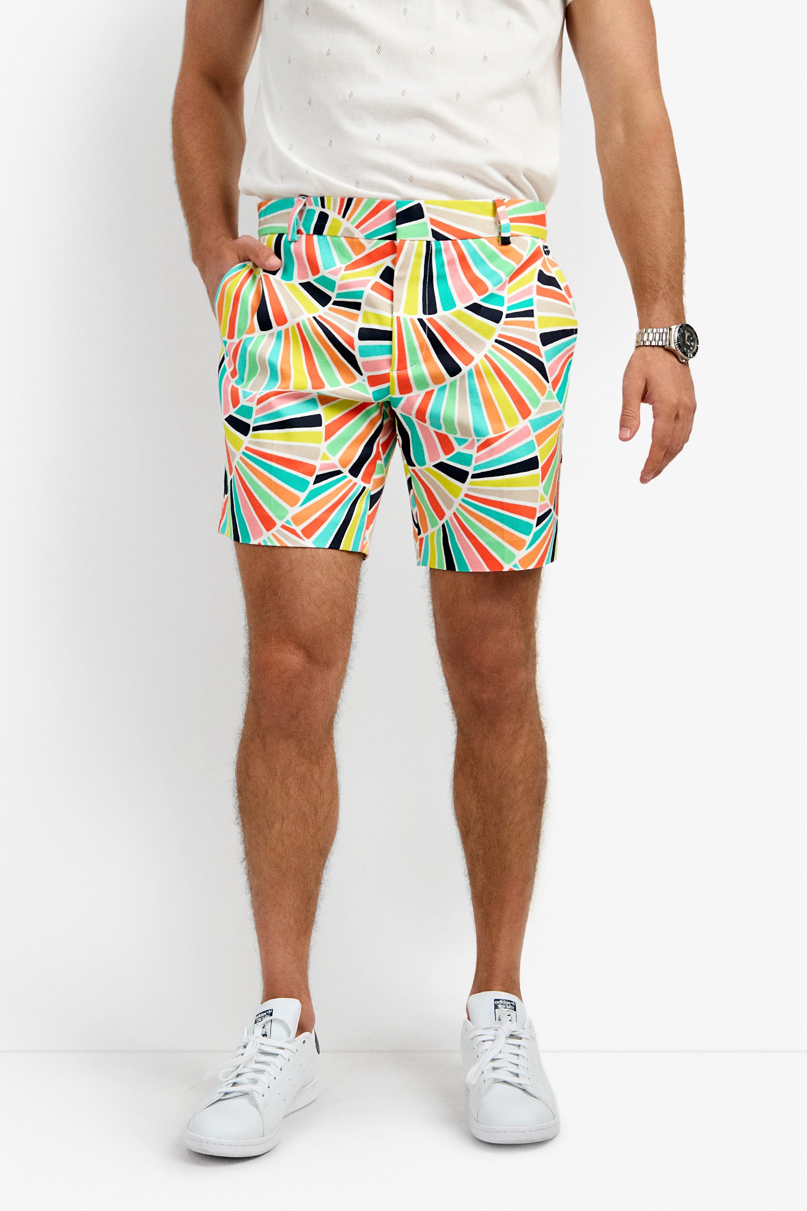 A person wears a white shirt, the Lawrence Short featuring colorful geometric patterns, white sneakers, and a watch, standing against a plain white background with hands in pockets.