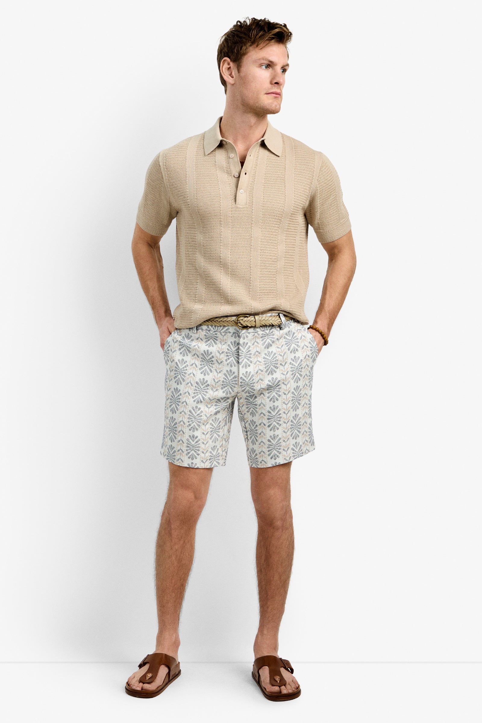 A man stands against a white background wearing the Lawrence Short, a beige knit short-sleeve polo, a braided belt, and brown sandals. His hands are in his pockets as he gazes to the side.
