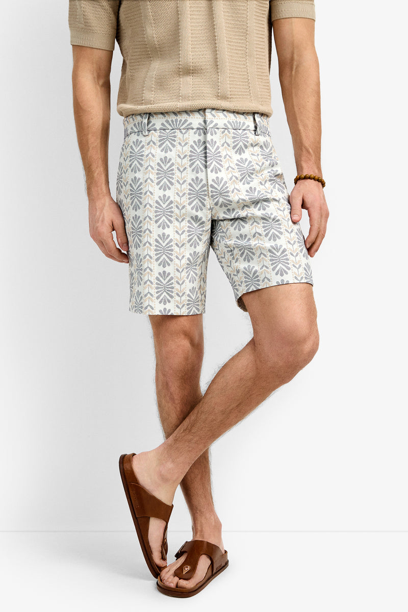 A man stands against a plain white background wearing a beige textured short-sleeve shirt, geometric patterned Lawrence Shorts in light hues, a beaded bracelet, and brown leather sandals.