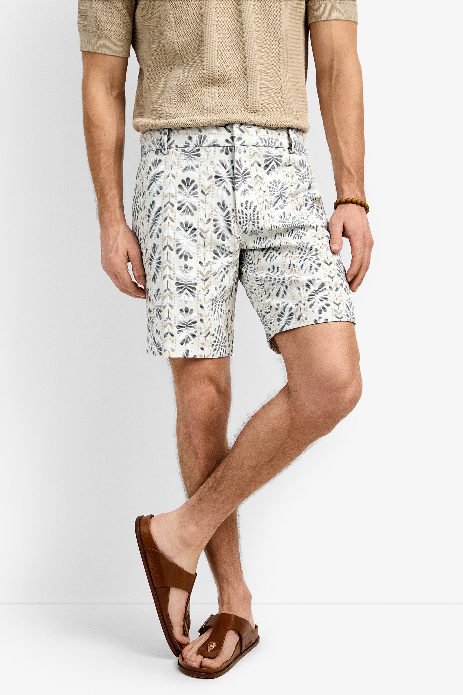 A man wearing the Lawrence Short, a beige short-sleeve knit shirt, brown sandals, and a beaded bracelet stands with legs crossed against a plain background.