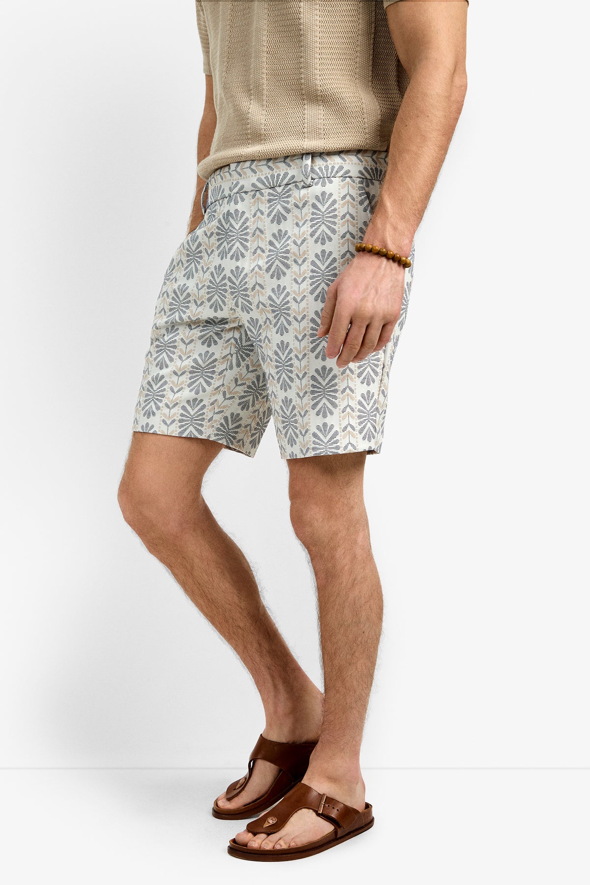 A man wearing the Lawrence Short, a beige short-sleeve knit shirt, brown sandals, and a beaded bracelet stands against a plain white background.