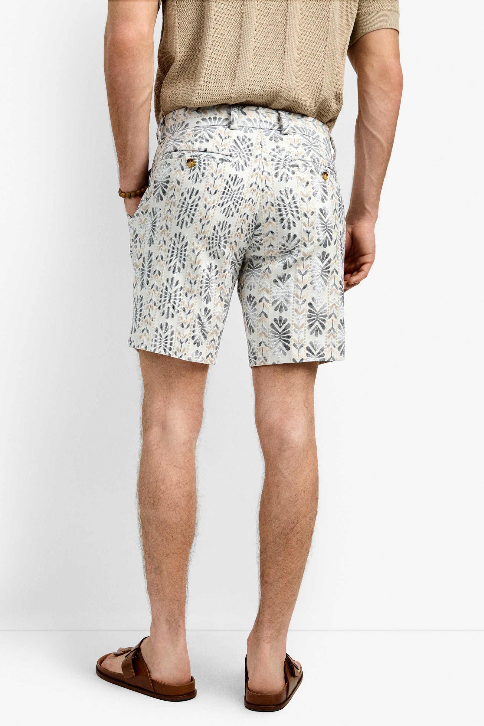 A man faces away, wearing the Lawrence Short with a beige and gray geometric pattern, a light brown short-sleeve knit shirt, brown sandals, and a bracelet on his left wrist.