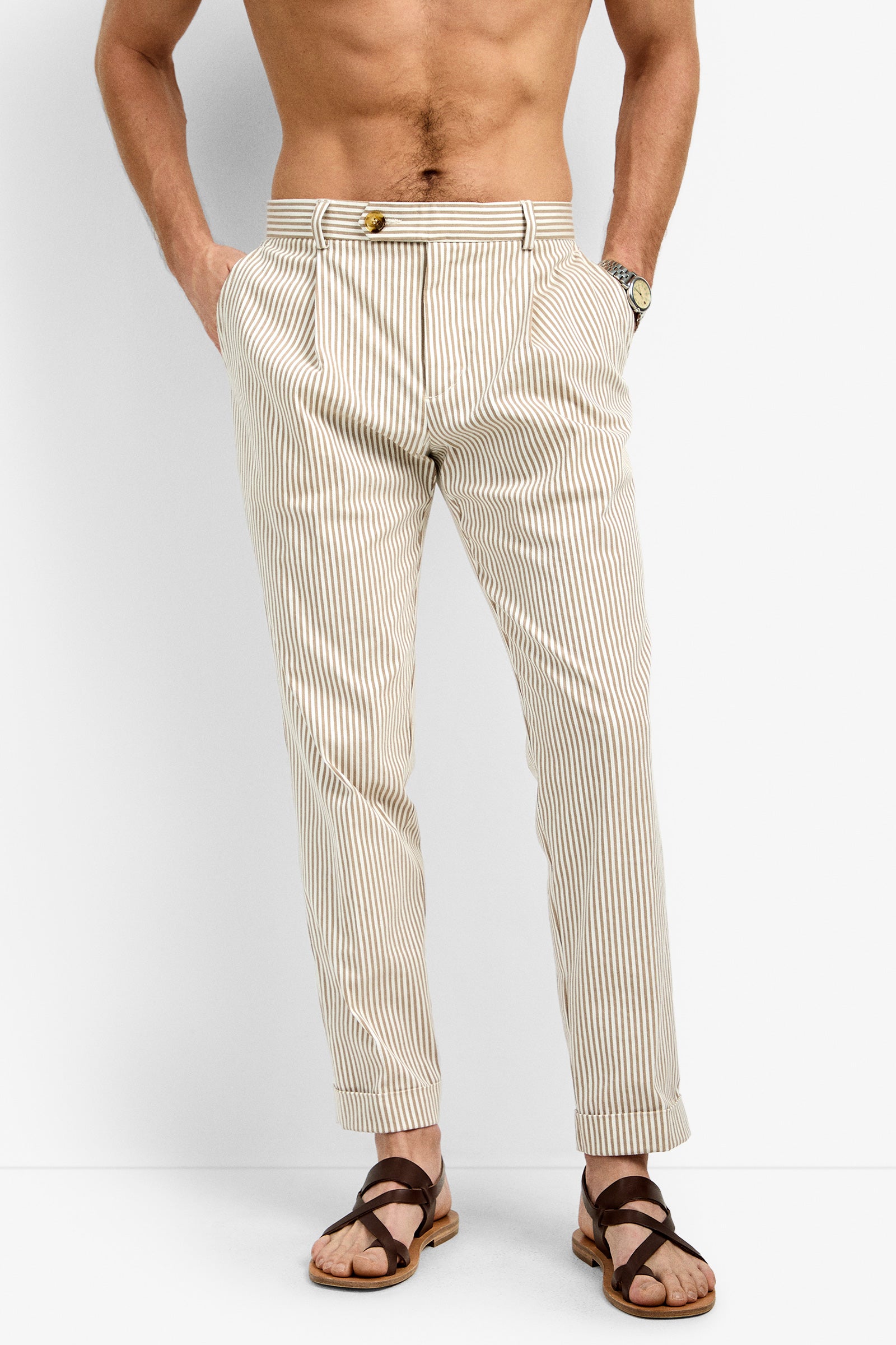 A shirtless man stands with his hands in his pockets, wearing Beal Pleated Trousers in Beachy Beige/White Wash and brown sandals against a plain white background.