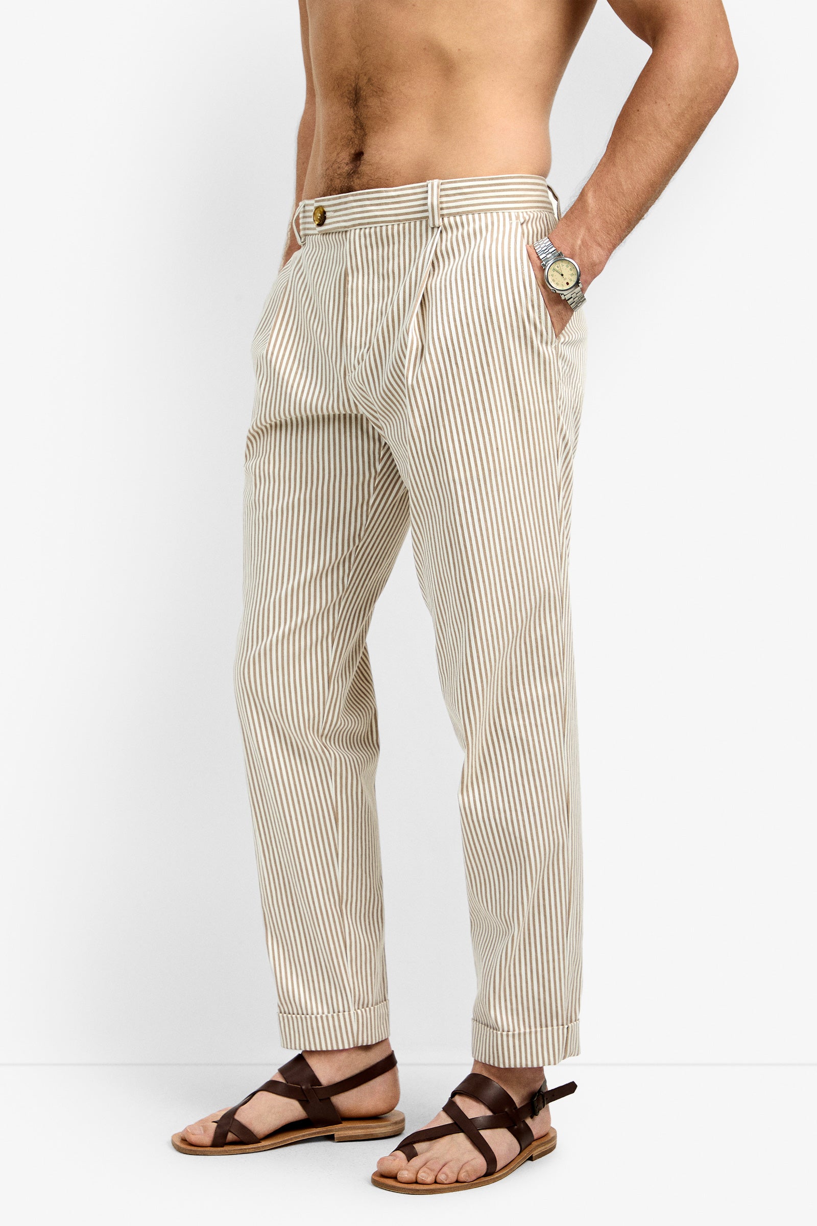 Wearing the Beale Pleated Trouser in beige and white vertical stripes, brown sandals, and a wristwatch, a person stands with hands in pockets against a plain white background. Only the lower body is clearly visible.