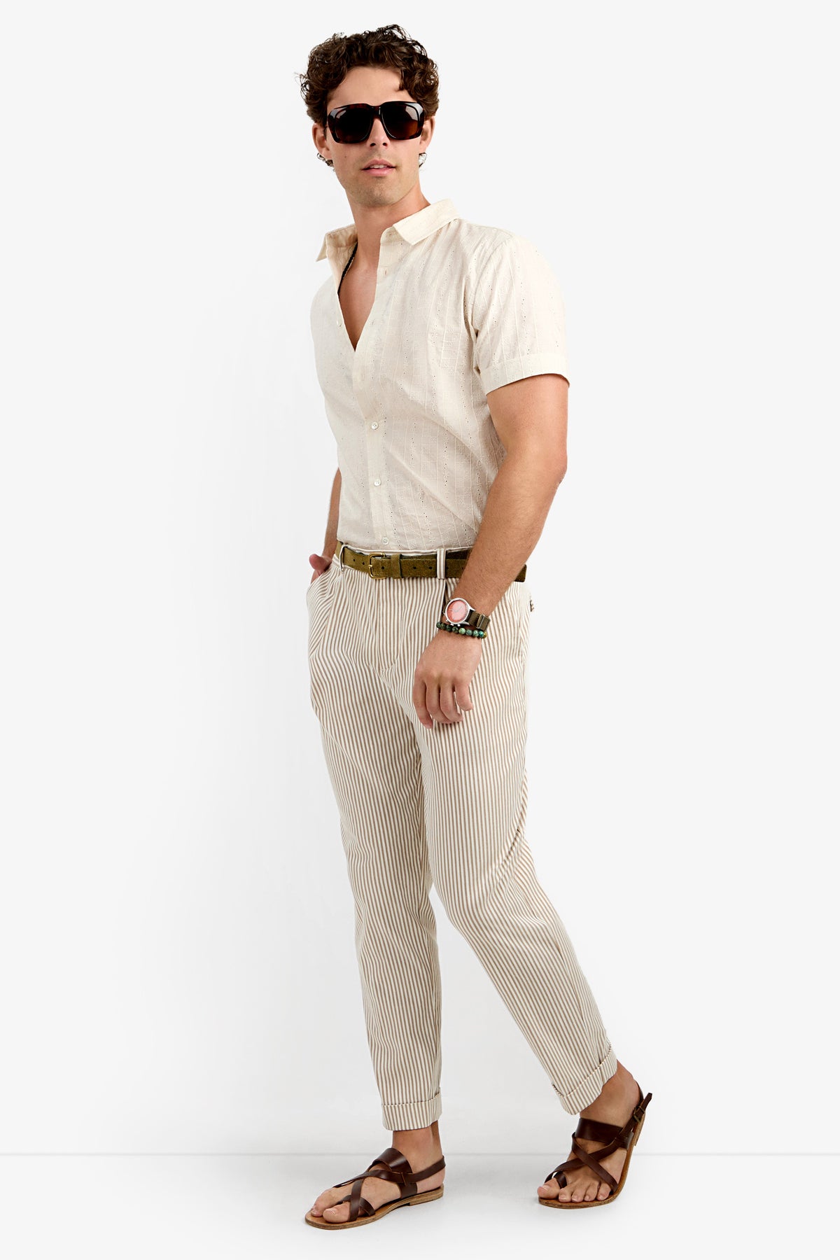A man stands confidently with one hand in his pocket, wearing sunglasses, a light short-sleeve shirt, a brown belt, brown sandals, a watch, and beige and white striped Beale Pleated Trousers against a plain white background.