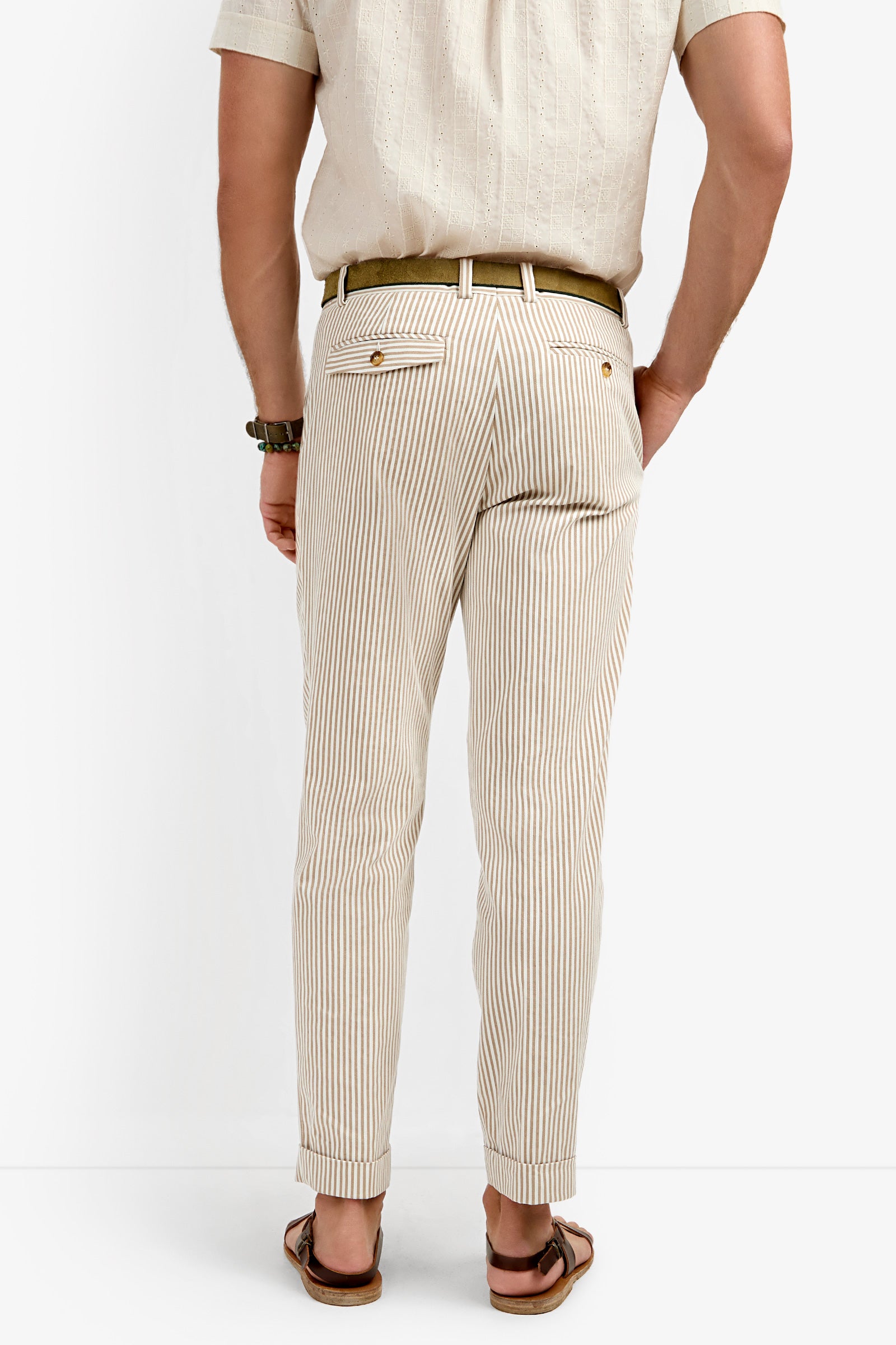 A man faces away, wearing Beale Pleated Trouser—light-colored with vertical stripes—paired with a beige short-sleeve shirt, brown belt, and sandals. His hands are by his sides against a plain white background.