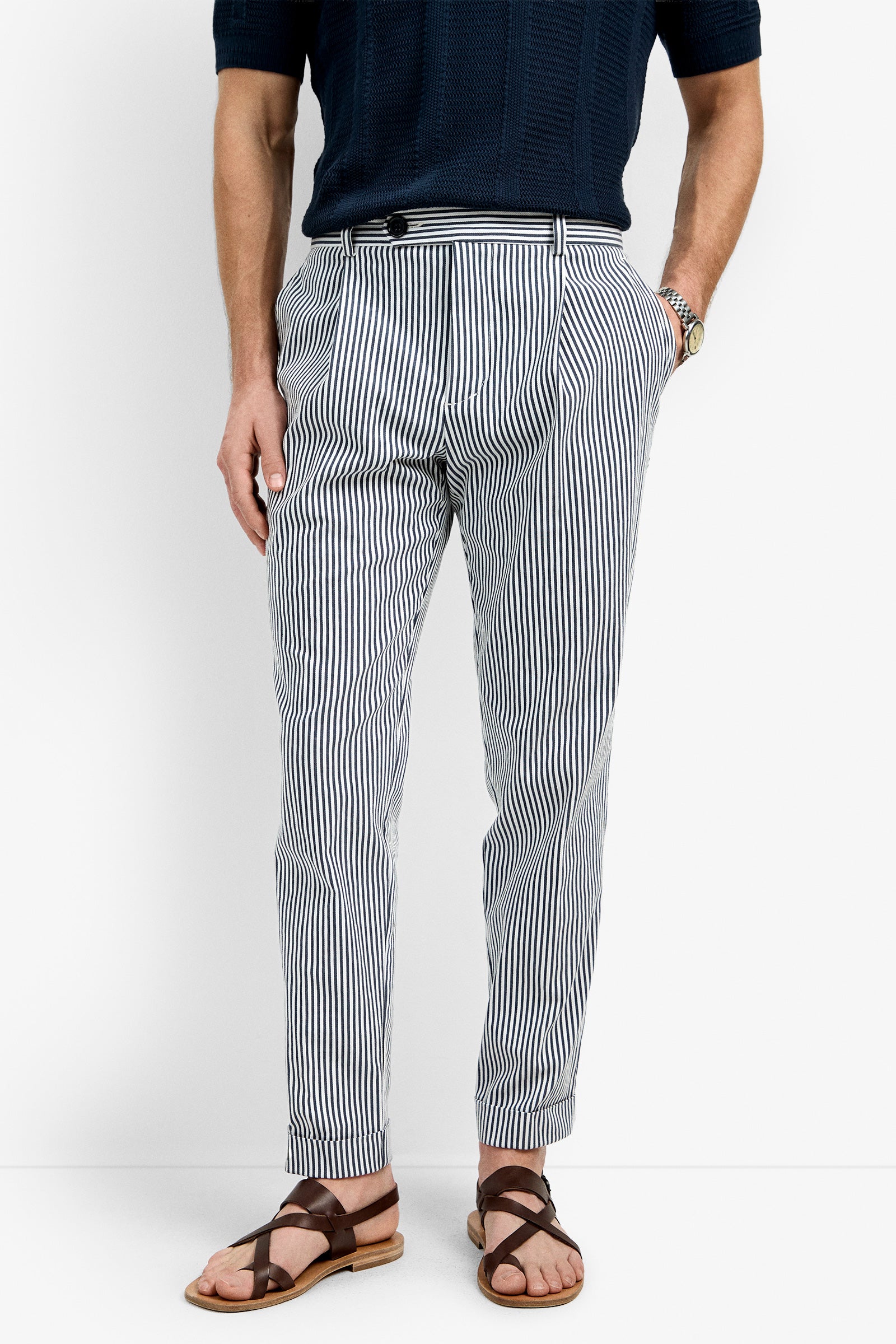 A man stands against a plain white background, hands in his pockets, wearing Beal Pleated Tourser in INK/WHITEWASH vertical stripes, a dark short-sleeve shirt, brown sandals, and a wristwatch.
