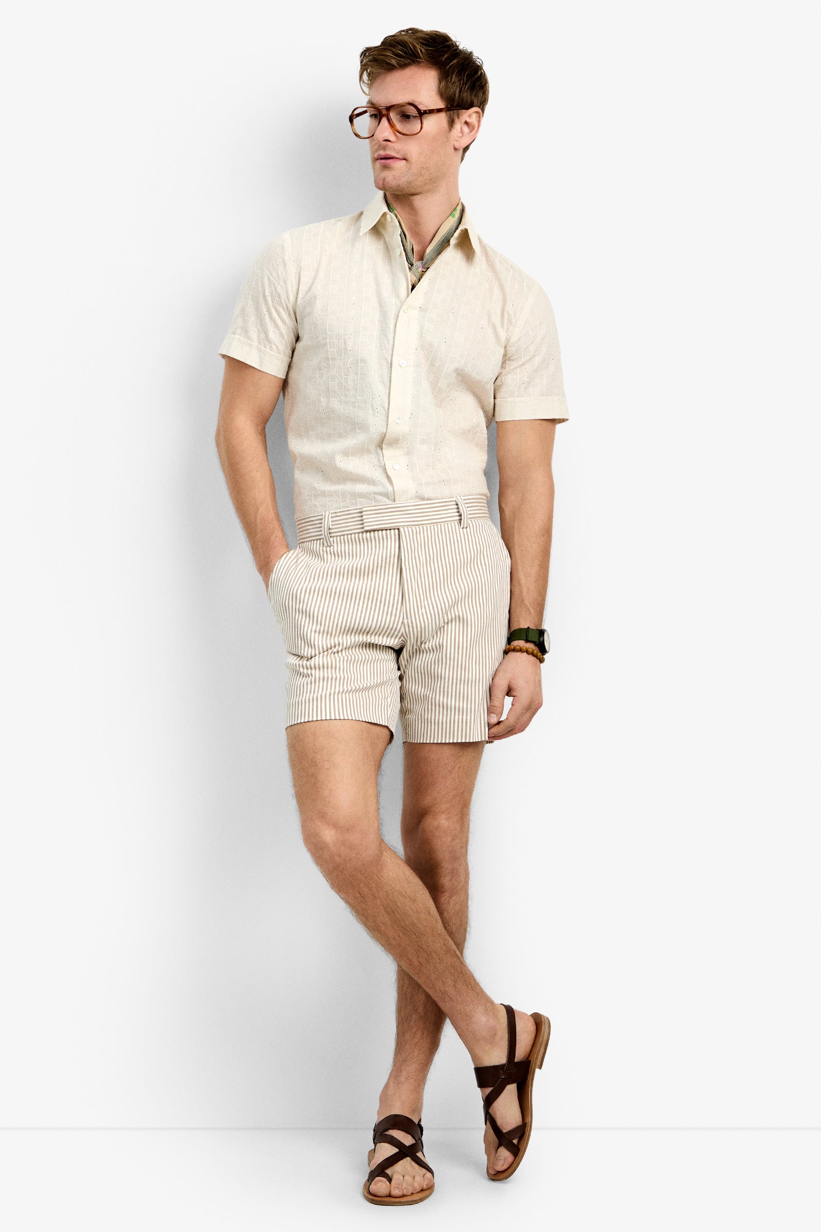 A man wearing glasses, a beige short-sleeve shirt, the Roger Short, brown sandals, and a watch stands with hands in pockets and one leg crossed in front of the other against a plain white background.