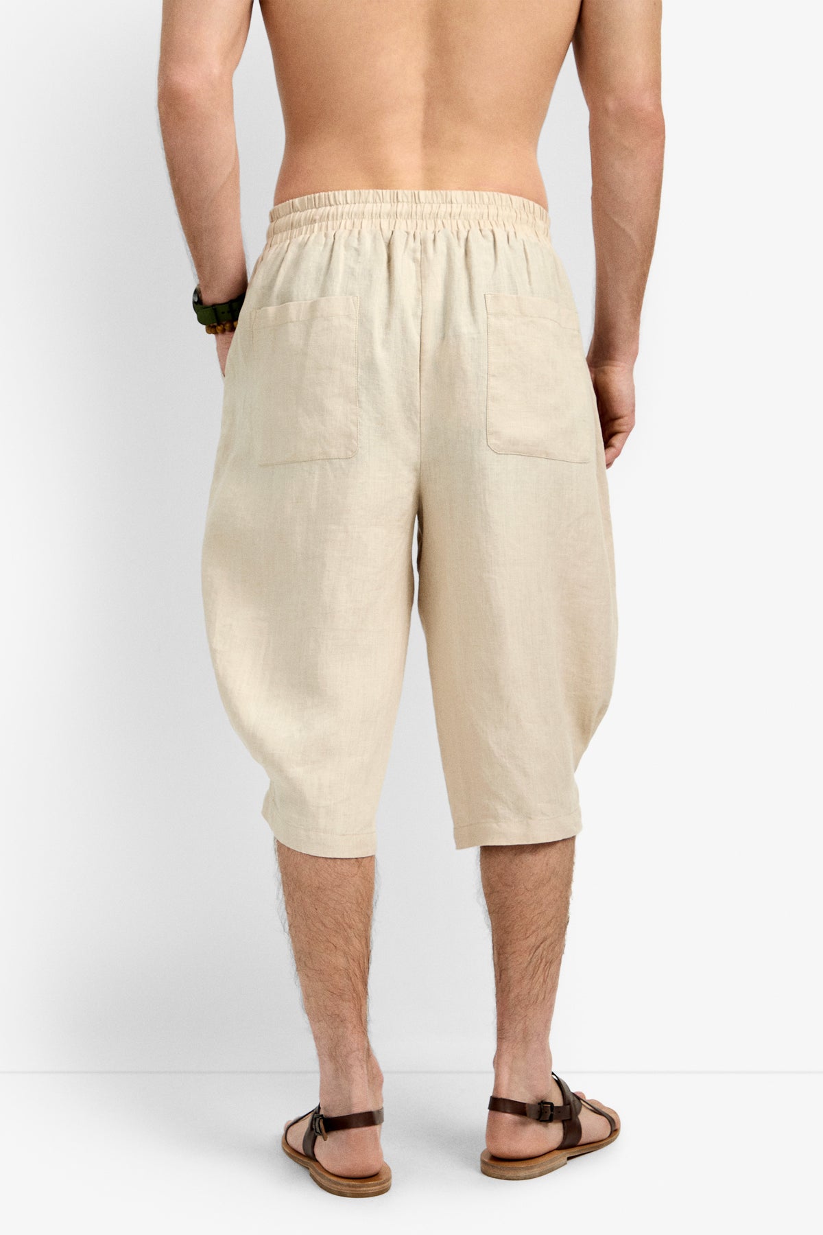 A shirtless man stands facing away, wearing the Claude Wide Leg Short in beige linen—loose, knee-length with an elastic waistband and two back pockets—paired with brown sandals and a wristwatch.