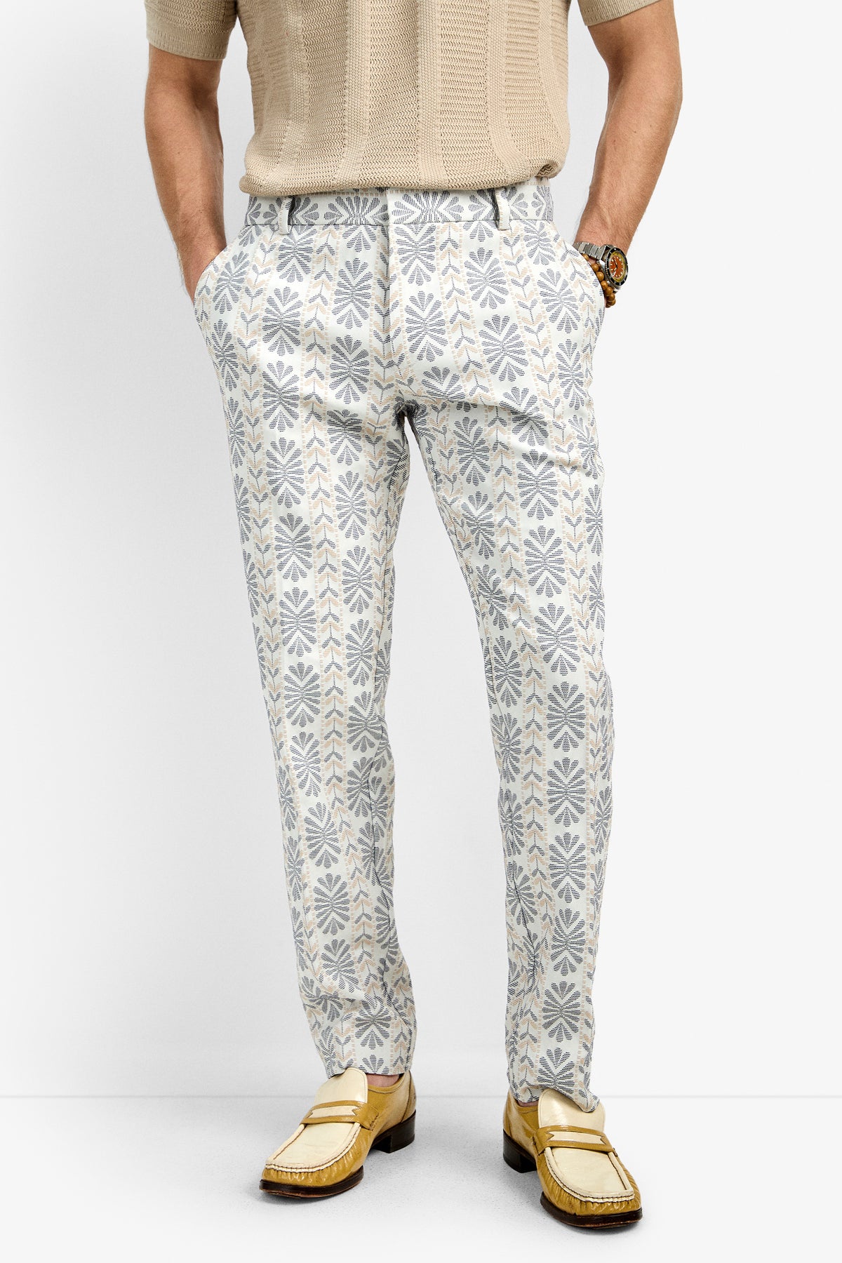 A person wearing Clyde Slim Trouser, which are patterned white and gray pants with a floral design, paired with a beige short-sleeve sweater and yellow loafers, standing hands in pockets against a plain white background.