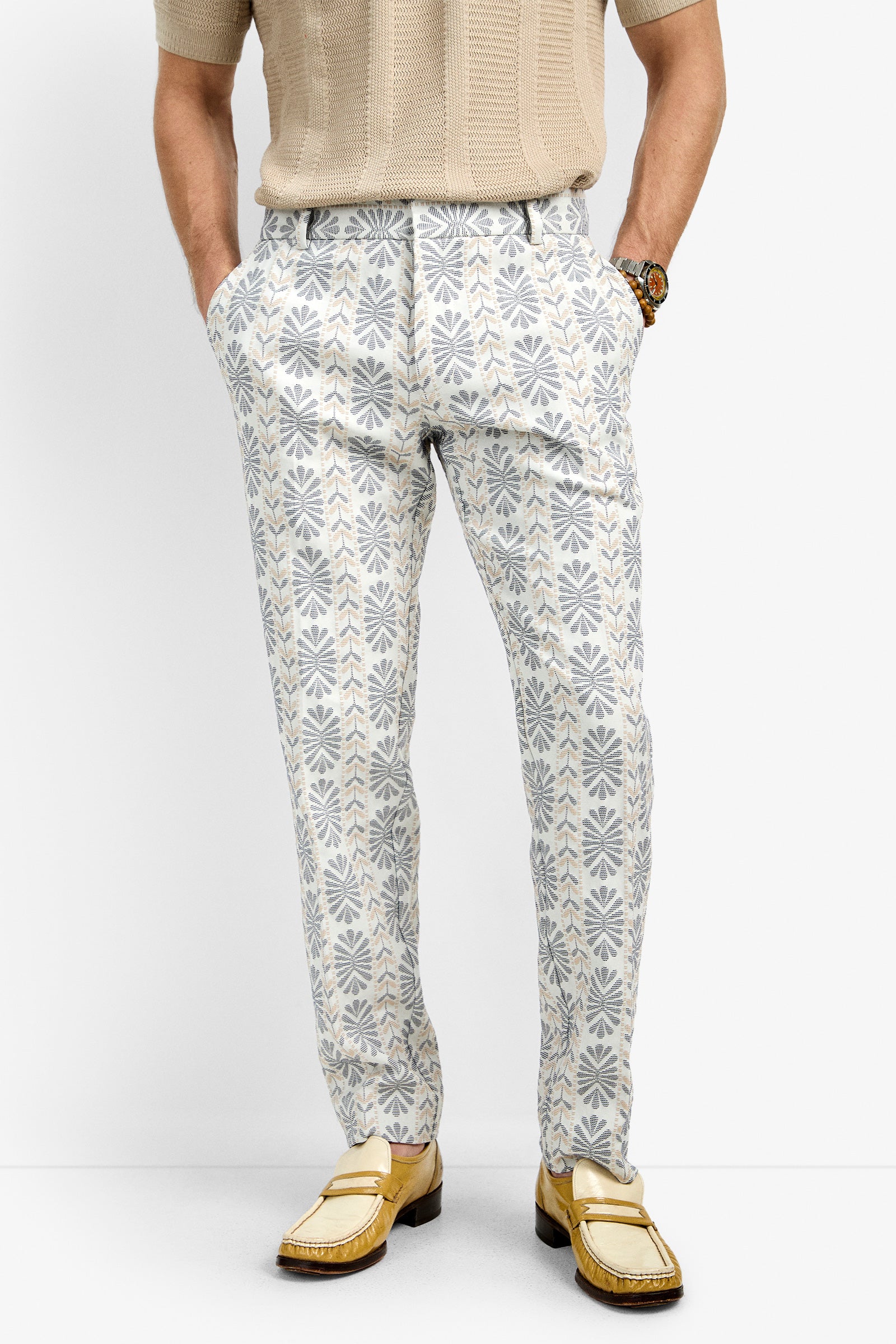 A person wearing Clyde Slim Trouser, which are patterned white and gray pants with a floral design, paired with a beige short-sleeve sweater and yellow loafers, standing hands in pockets against a plain white background.