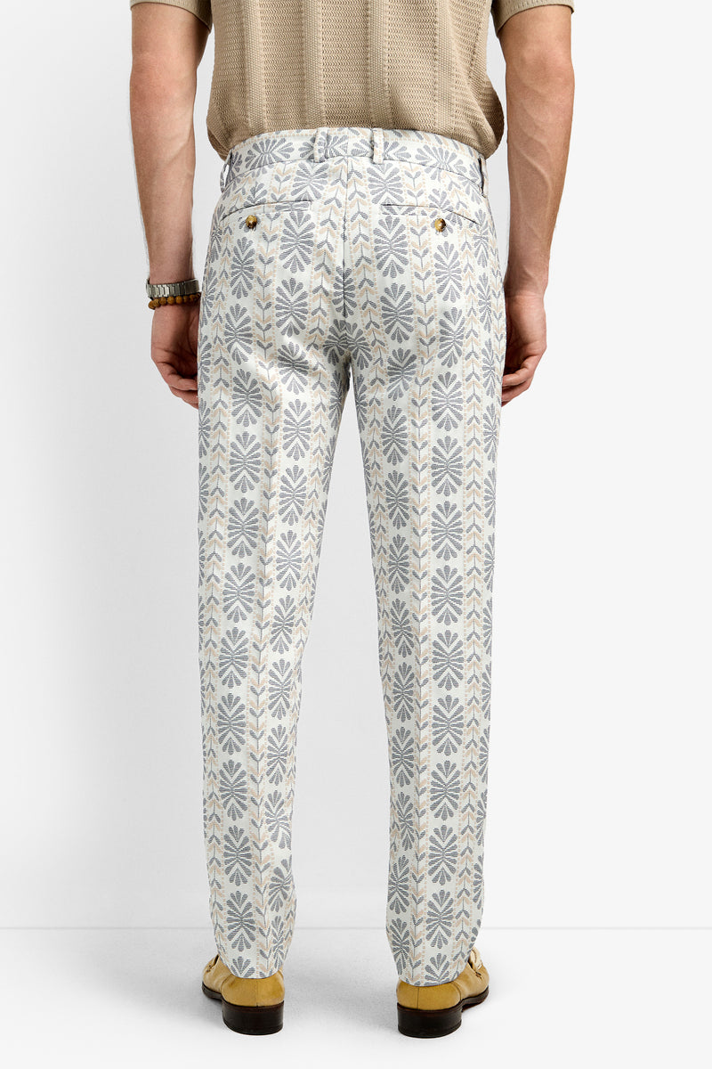 A person stands facing away against a plain white background, wearing the Clyde Slim Trouser in a blue and beige geometric floral pattern, paired with a tan short-sleeve shirt, watch, and tan loafers.
