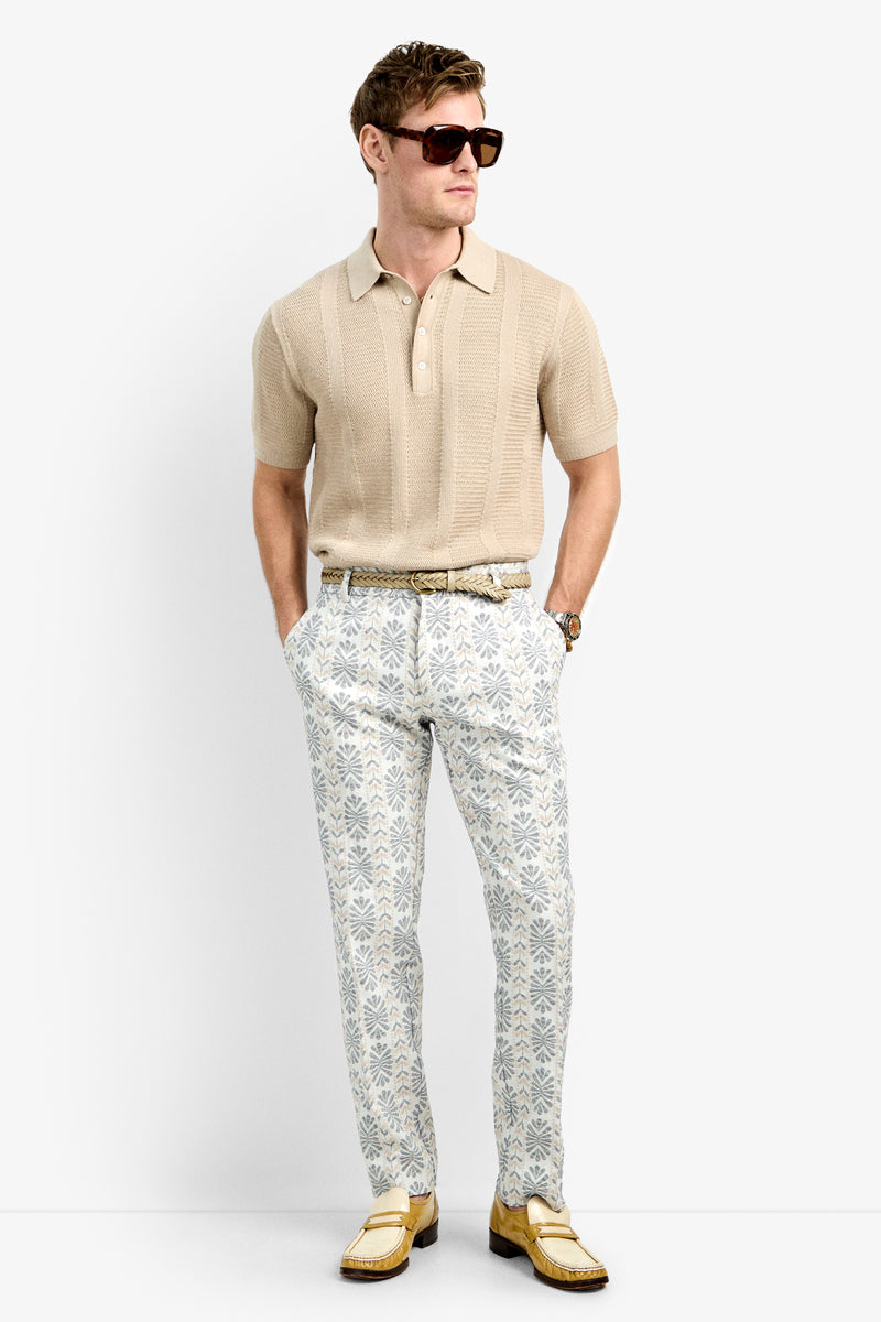 A man wearing a beige short-sleeve polo, Clyde Slim Trousers in patterned white and gray, yellow loafers, sunglasses, and a watch stands with his hands in his pockets against a plain white background.