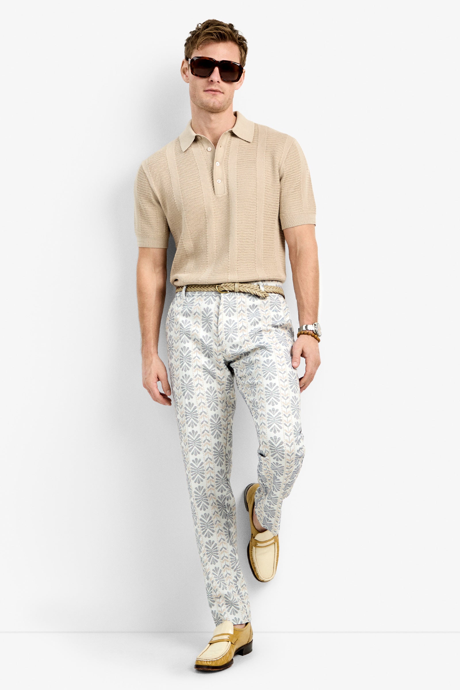 A man wearing sunglasses, a beige short-sleeve polo shirt, Clyde Slim Trousers in white and blue pattern, a belt, and beige loafers stands against a white background.
