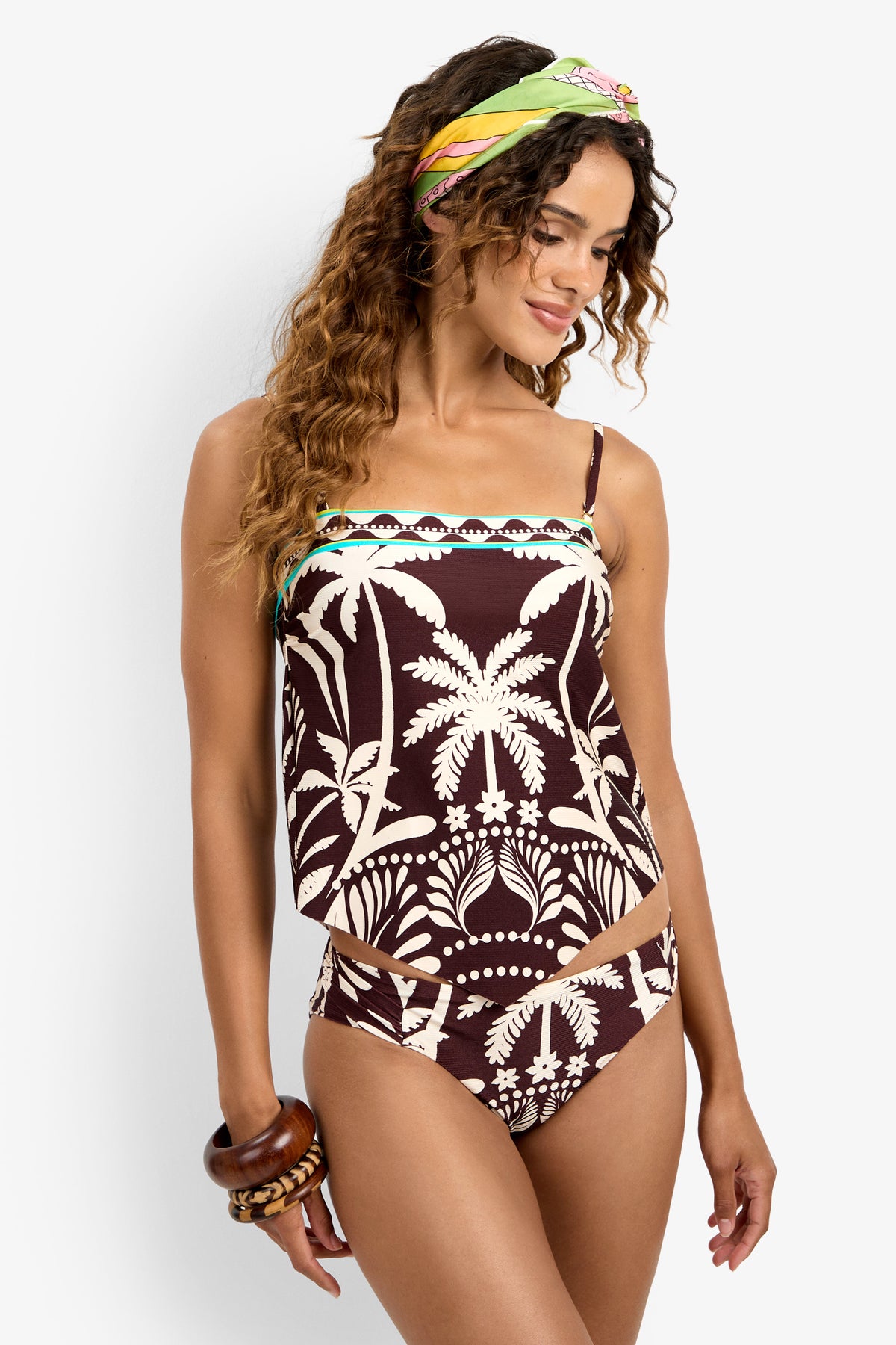 Woman models the Handkerchief Tankini Swim Top with a brown and beige tropical palm print, styled with a vibrant headscarf and chunky bracelets, smiling against a white background.