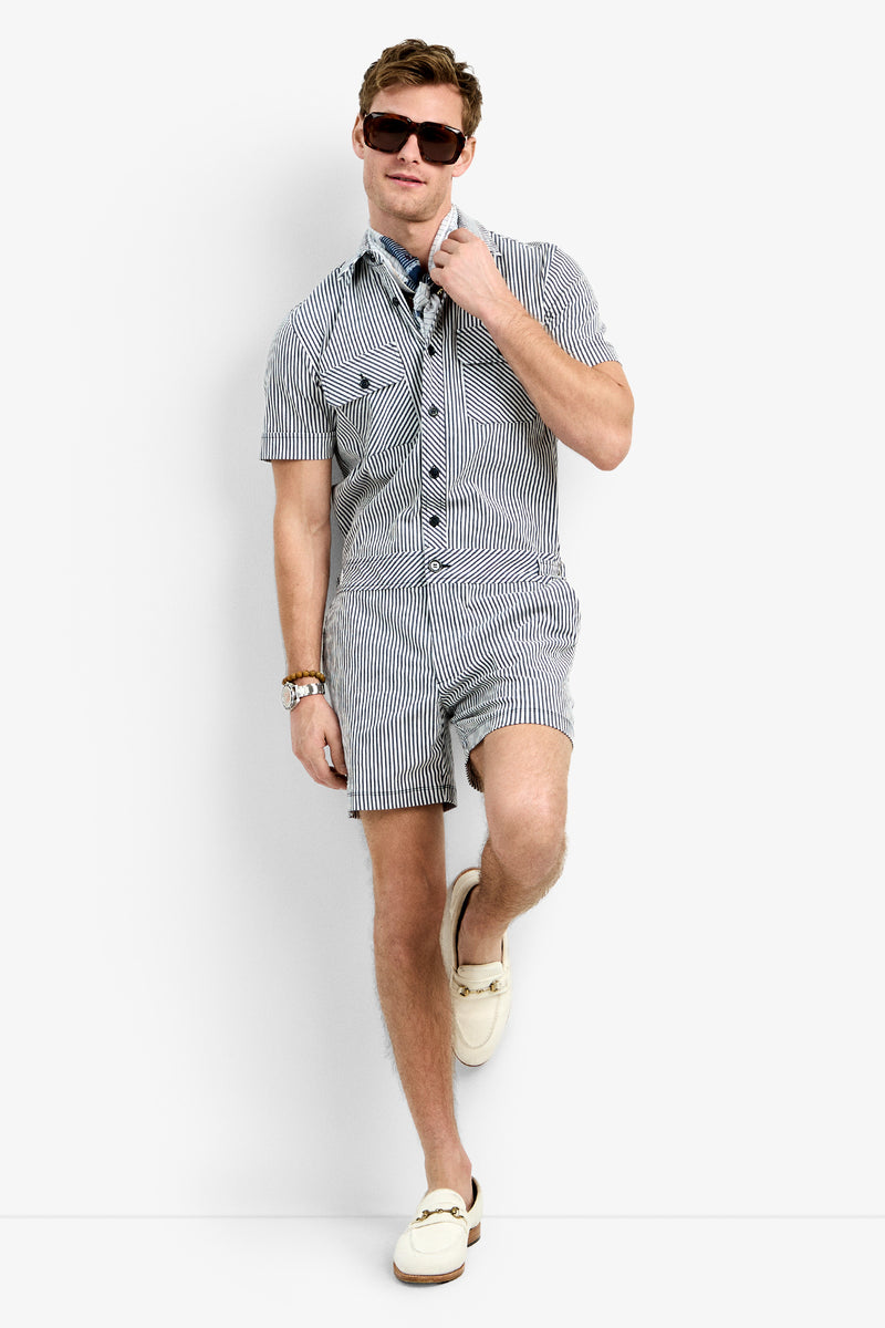 A man in the Sherman Short Jumpsuit, sunglasses, a watch, a blue patterned scarf, and cream loafers stands against a white background, leaning with one leg crossed.